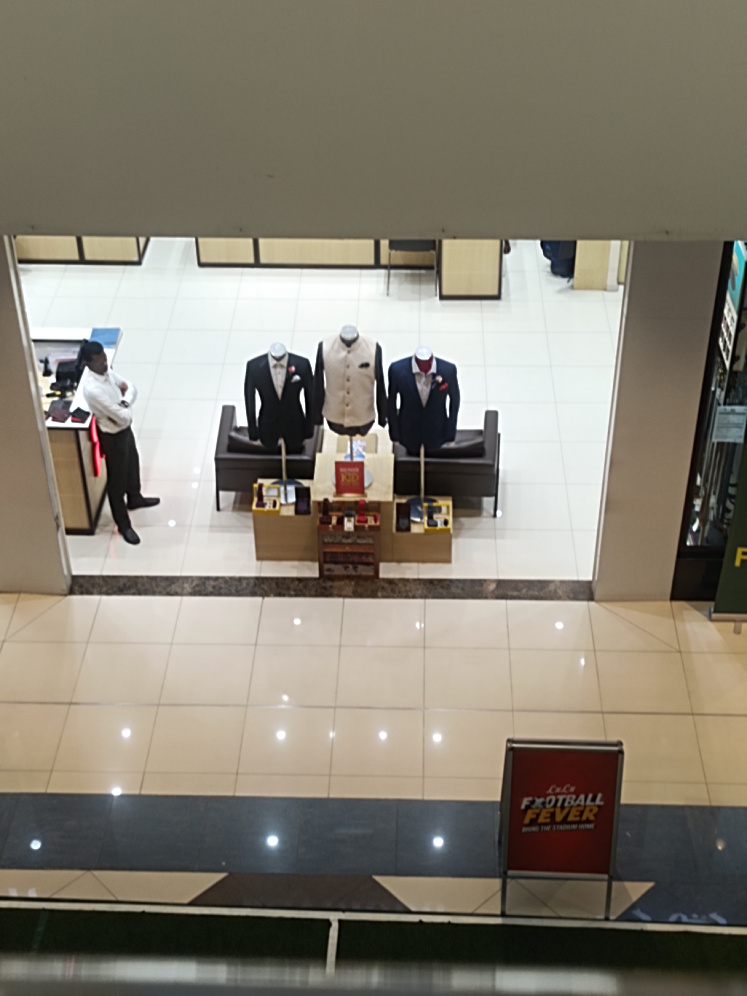An aerial view captures a section of a retail store, likely a men's clothing establishment, in Dubai, United Arab Emirates. The scene is dominated by polished tile flooring and white walls. In the mid-ground, three mannequins are displayed: one wearing a dark suit with a white shirt and a red pocket square; another in a light-colored waistcoat over a white shirt; and a third in a dark blue suit. A dark brown sofa sits behind the mannequins. To the left, a male store employee, wearing a white long-sleeved shirt and black trousers, stands with his arms crossed, facing away from the camera. Adjacent to him is a counter area with various items displayed. Further in the foreground, a red promotional sign with the text "LuLu FOOTBALL FEVER BRING THE STADIUM HOME" is visible. The overall impression is of a well-lit, modern commercial space.