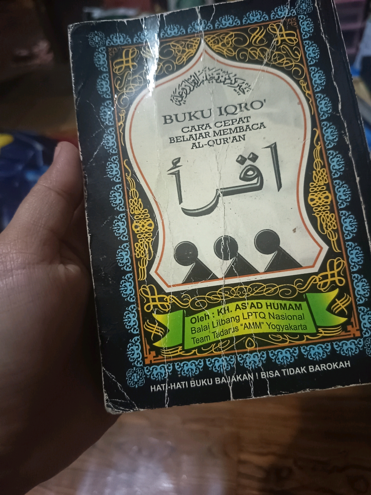 A book titled "BUKU IQRO' CARA CEPAT BELAJAR MEMBACA AL-QUR'AN" is held in a person's left hand. The book has a dark blue cover with intricate gold and light blue Islamic-inspired patterns along the edges. The center of the cover features the title in white and orange text, with a large Arabic word "اقرأ" (iqra') in black calligraphy. Below this, there is a green banner with white text stating "Oleh: KH. AS'AD HUMAM Balai Litbang LPTQ Nasional Team Tadarus "AMM" Yogyakarta". At the very bottom of the cover, in white text, it reads "HATI-HATI BUKU BAJAKAN! BISA TIDAK BAROKAH". The background is a blurry wooden surface. The image is taken from a slightly elevated angle, showing the book being held in the foreground. The lighting is natural and somewhat dim. The location is not identifiable from the image itself.