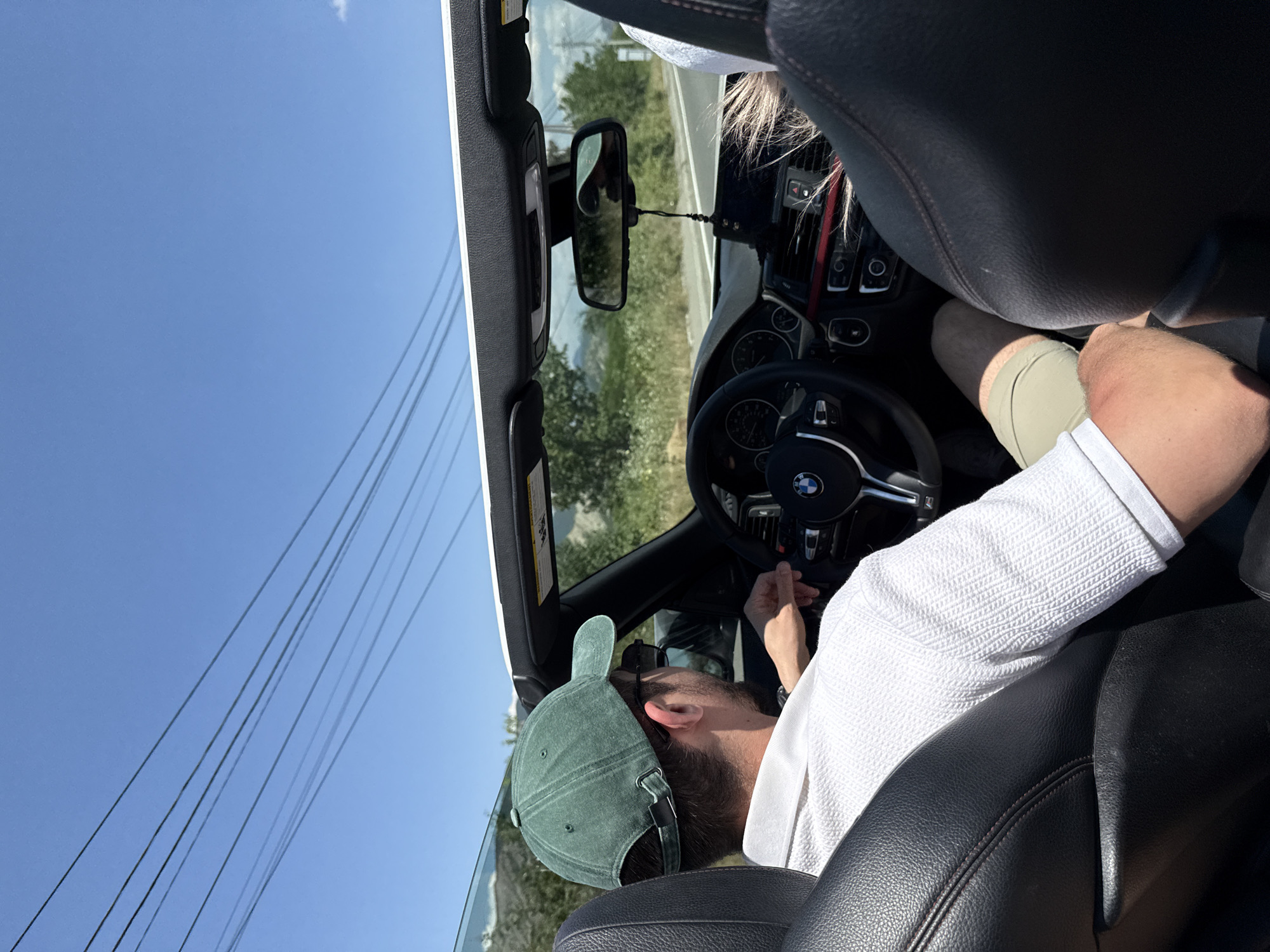 The image shows a view from the back seat of a convertible car. The primary subject is a person wearing a green baseball cap and a white shirt, seated in the driver's seat. They appear to be reaching for something on the dashboard. Sunlight streams in, indicating it is daytime. The car's interior is visible, including the black leather seats, steering wheel with a BMW logo, and part of the dashboard. Outside the car, a clear blue sky is visible with some power lines in the foreground. The background shows a rural landscape with trees and hills under the bright sky, suggesting a sunny day in a potentially rural or semi-rural location. The overall mood is casual and relaxed, consistent with a scenic drive on a pleasant day. No visible text is present in the image.