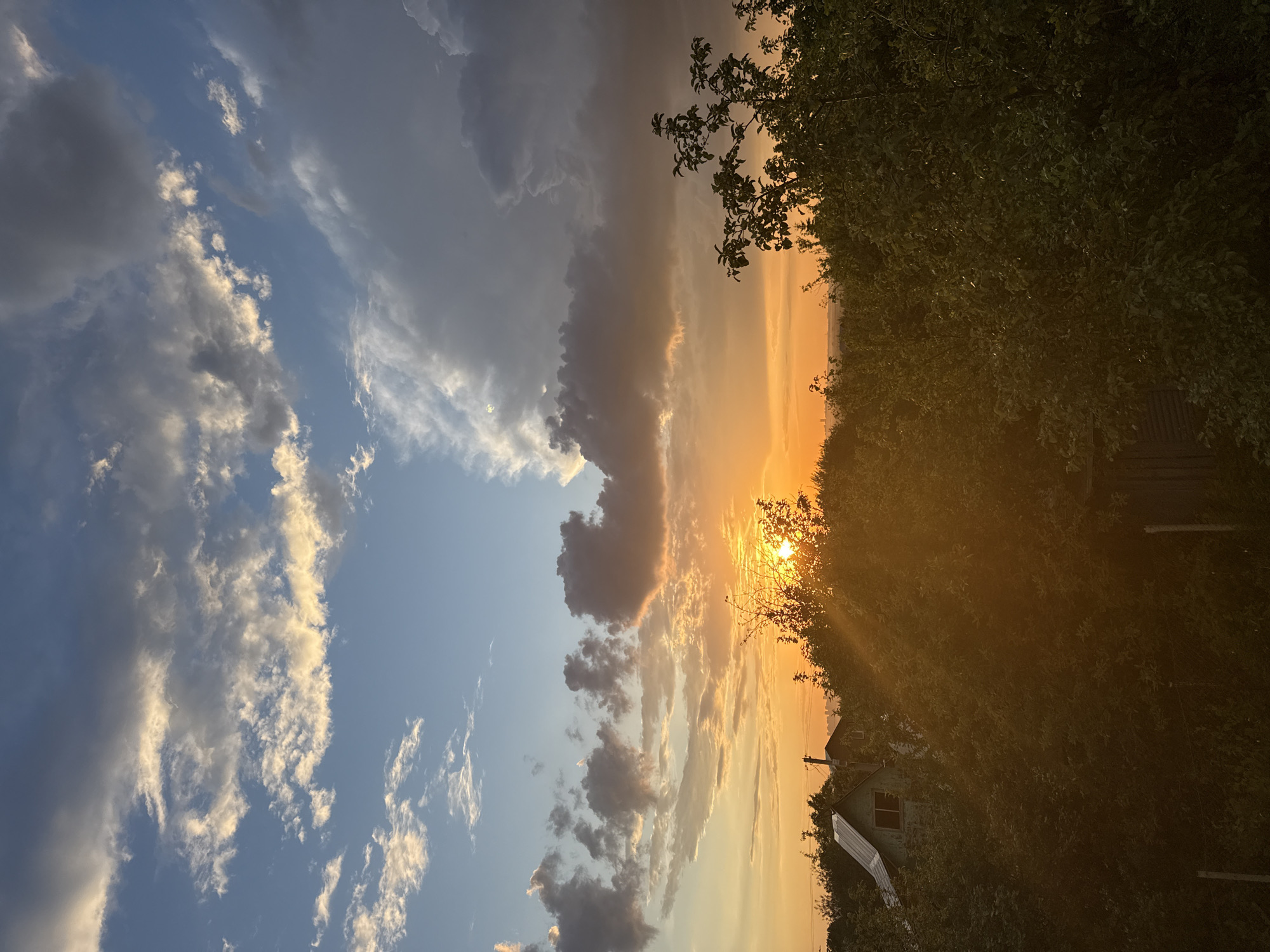 The image depicts a sunset over a semi-rural area. The sun is low on the horizon, casting a warm orange glow and creating sunbeams that filter through the foliage. In the foreground, there is dense green foliage, with the tops of trees and bushes dominating the lower portion of the frame. A small, light-colored house with a dark roof is partially visible amidst the greenery on the left side of the image. Further in the background, other structures and trees are visible, appearing as silhouettes against the bright sky. The sky is a dramatic canvas of clouds; in the upper portion, there are wispy white and gray clouds against a blue sky, while closer to the horizon, larger, darker clouds are illuminated by the setting sun, creating a contrast of light and shadow. The overall scene conveys a sense of a peaceful evening in a suburban or rural setting. There are no people visible in the image.