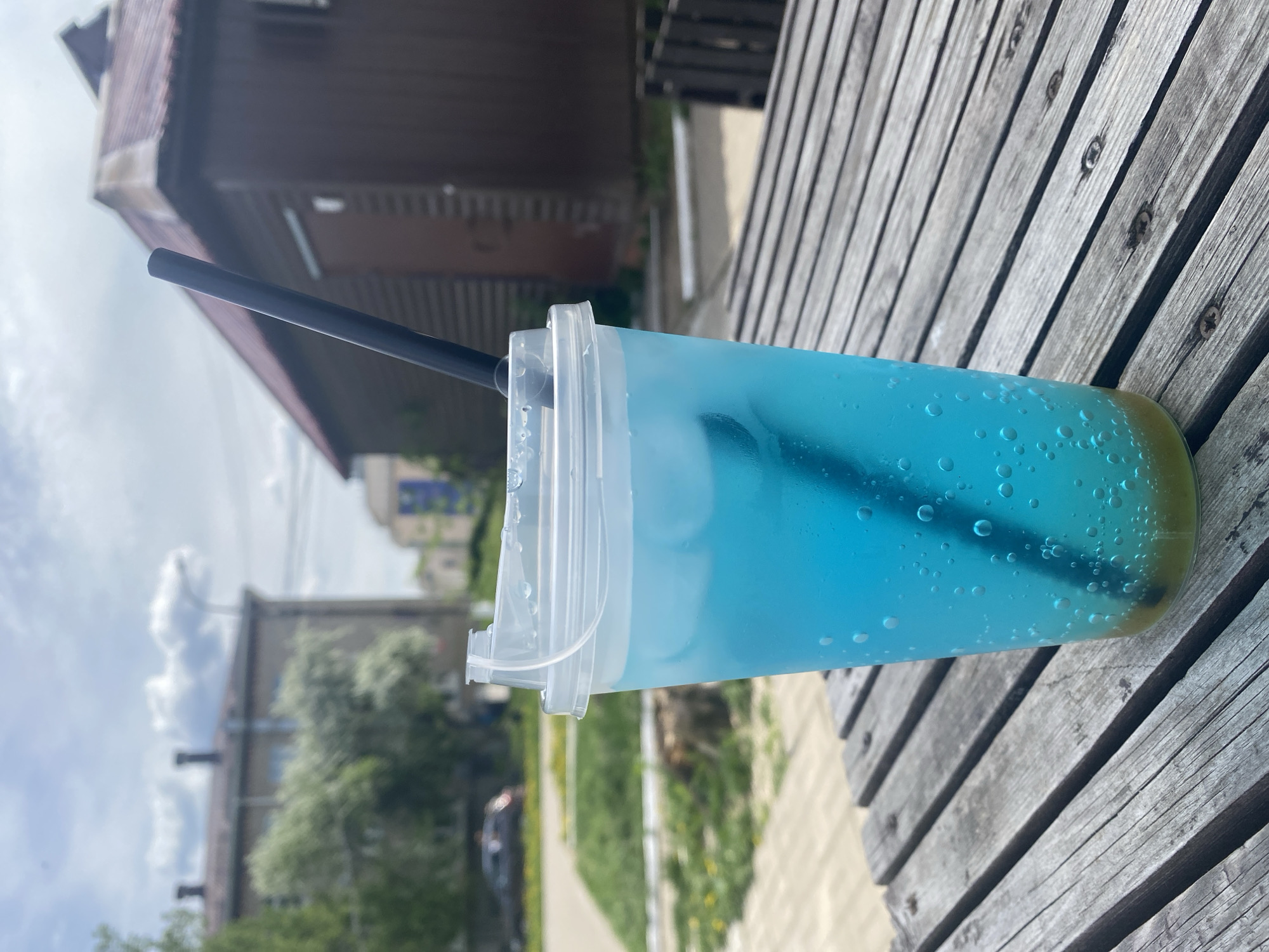 A plastic cup containing a blue liquid and ice sits on a wooden surface, presumably a bench. A black straw is inserted into the cup. The liquid has small bubbles throughout and a yellowish layer at the bottom. The cup has a clear lid with a slightly raised edge.
In the background, there are buildings and greenery, partially obscured by the cup. The image is taken outdoors in daylight. The buildings appear to be urban structures with a mix of residential and commercial designs. The sky is partially visible with clouds. The bench is made of wooden planks with visible grain and wear.
The location cannot be determined with certainty to be the city of Chaikovsky, Russia without further information.