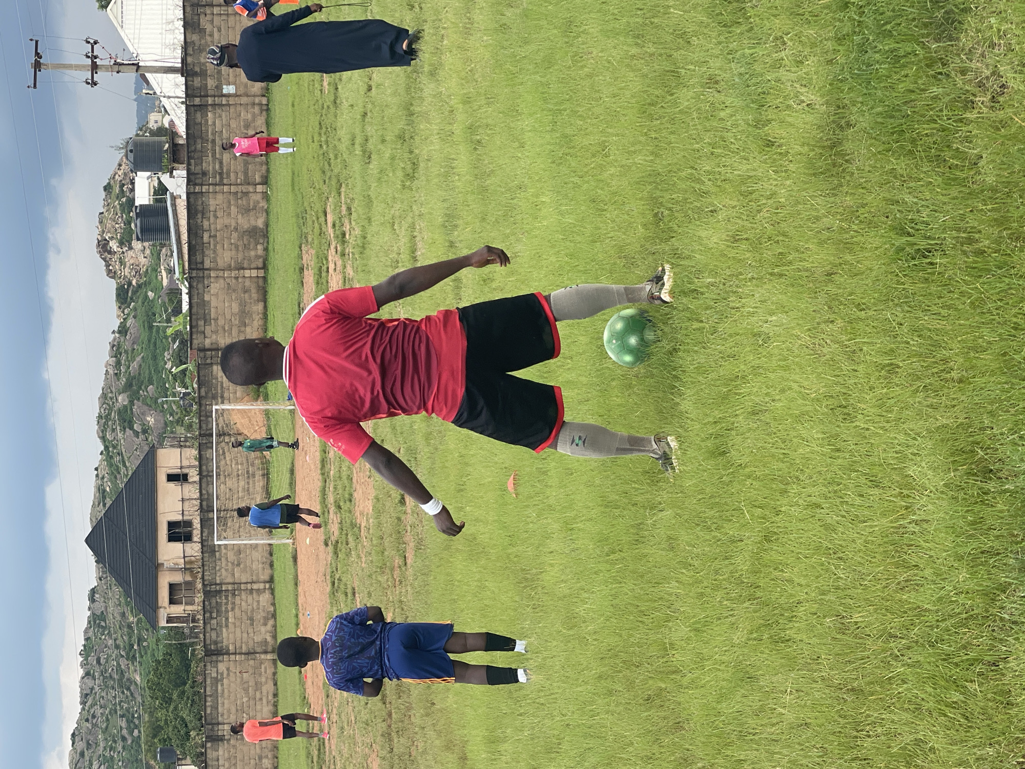 An outdoor, low-angle, medium shot captures a group of young men engaged in a soccer training session on a grassy field. The scene is set in Jos, Nigeria, with a backdrop of rocky hills and some residential buildings. The sky is overcast, suggesting a cloudy day.
In the foreground, a young man wearing a red t-shirt and black shorts with red trim is facing away from the camera, positioned behind a green soccer ball. He has a white wristband on his left arm and is wearing grey socks and black and white soccer cleats. To his left and slightly behind him, another young man is seen from the back, wearing a blue t-shirt with a dark pattern and blue shorts with yellow stripes, also with black socks and white soccer cleats.
In the middle ground, a soccer goal with a white net stands on a patch of exposed dirt. Several other individuals are visible near the goal. One person in an orange t-shirt and black shorts is facing away from the camera, standing to the left of the goal. Two other figures, one in a blue vest and black shorts and another in a green t-shirt and black shorts, are closer to the goal. Further to the right, a person in a pink t-shirt and white shorts stands facing the goal. A man in a dark, flowing robe and a cap, possibly a coach or observer, is positioned on the right side of the frame, further back on the field, holding what appears to be a lanyard or strap in his right hand.
The field itself is predominantly covered in lush green grass, with patches of bare earth around the goal area. Several orange cones are scattered on the field, indicating training drills. The environment suggests a community or school sports ground. The overall atmosphere is one of active participation in a sporting event.