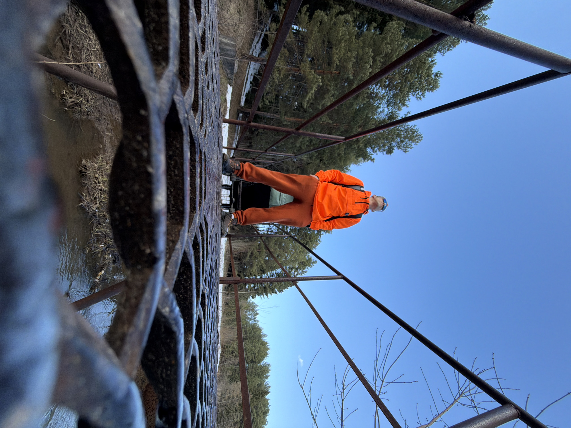 A low-angle, rotated view captures a person standing on a metal grate bridge. The person is wearing orange pants and a matching jacket. The bridge appears to be a pedestrian walkway spanning a small body of water, likely a stream or river, with muddy banks visible on the left side of the image. Trees line the background, and the sky is clear and blue. The location is the city of Chaikovsky, Russia.