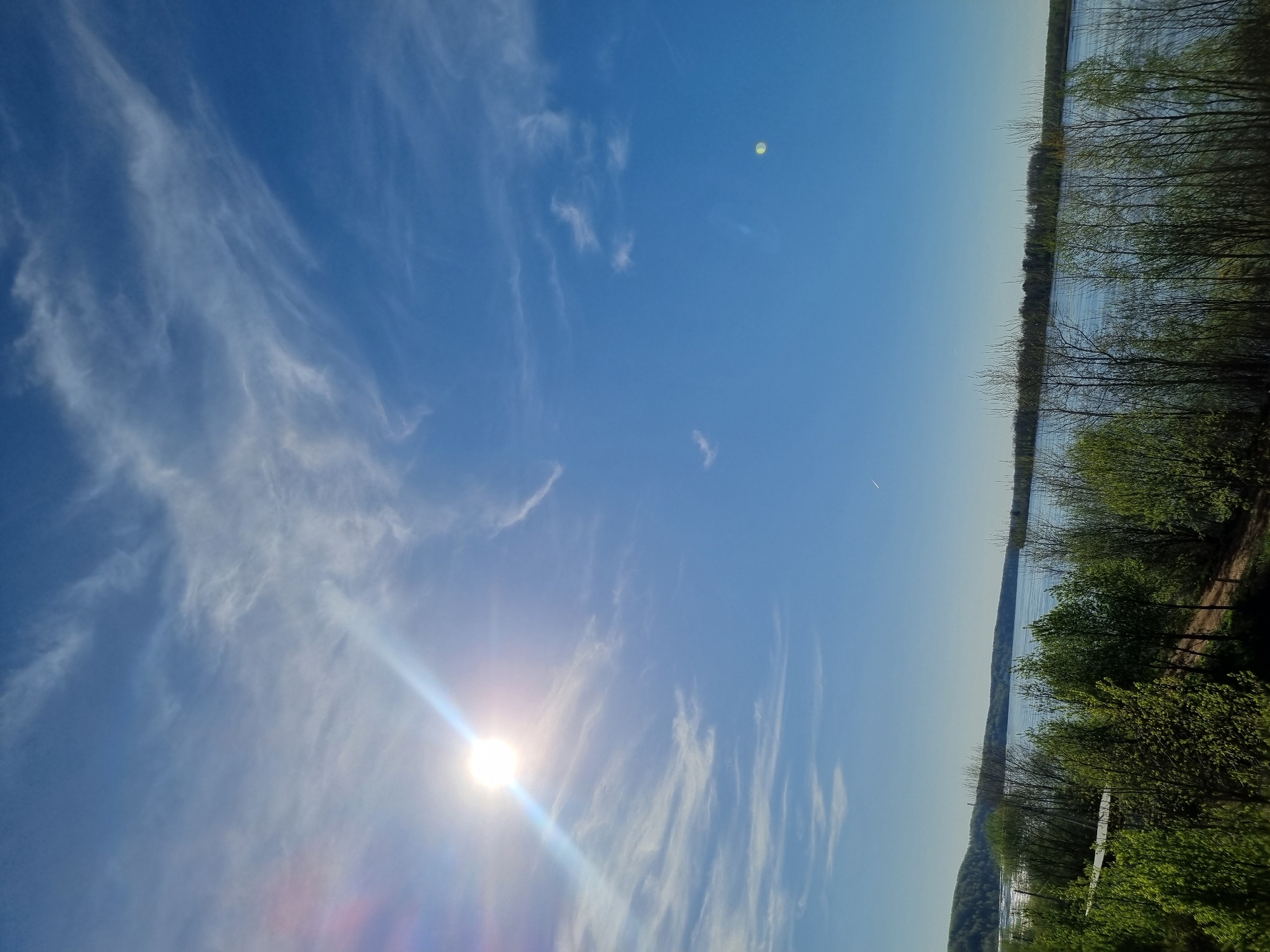 The image depicts a daytime scene in Ольховка, Russia, with a bright blue sky and wispy cirrus clouds. The sun is visible in the lower left corner of the frame, creating lens flare. A body of water, likely a river or lake, stretches horizontally across the middle ground, bordered on the right by trees and vegetation. The trees appear to be a mix of deciduous and coniferous types. A sliver of land is visible in the lower left, with hints of buildings. The overall impression is a clear, sunny day near a body of water, with signs of human habitation in the distance.