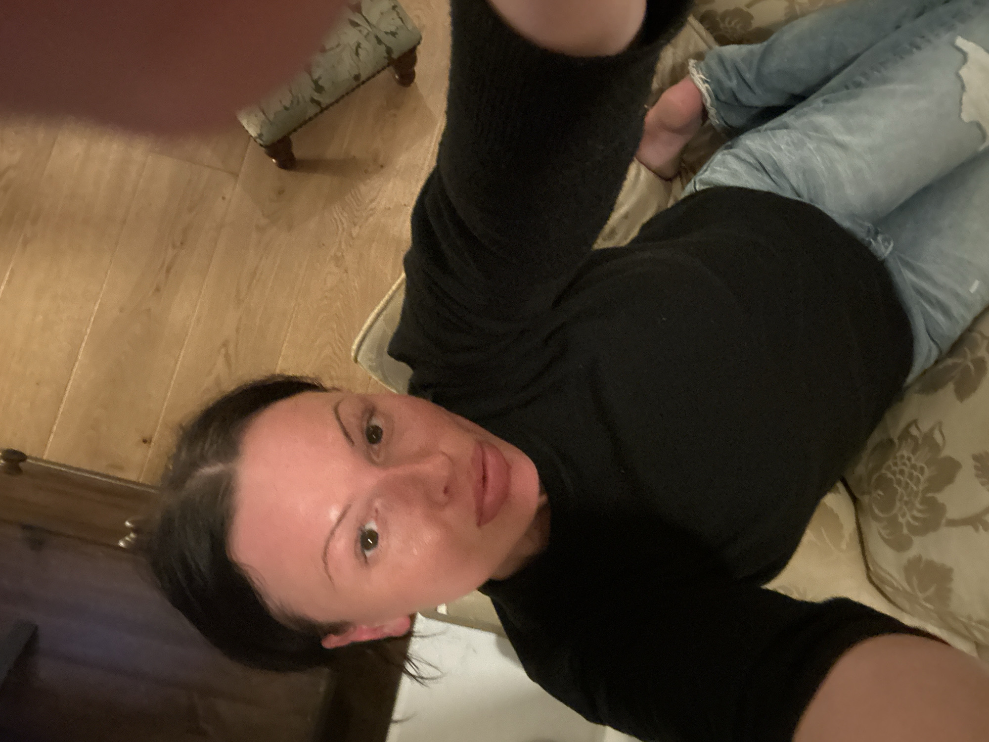 A selfie taken indoors shows a woman with dark hair pulled back from her face, wearing a black long-sleeved top and light-wash denim jeans. She is lying on a cream-colored sofa with a floral pattern. The room has wooden flooring, and a small upholstered stool with floral fabric is visible in the background. A dark wood cabinet is also partially visible. The location is likely within a home in Folkestone and Hythe District, United Kingdom.