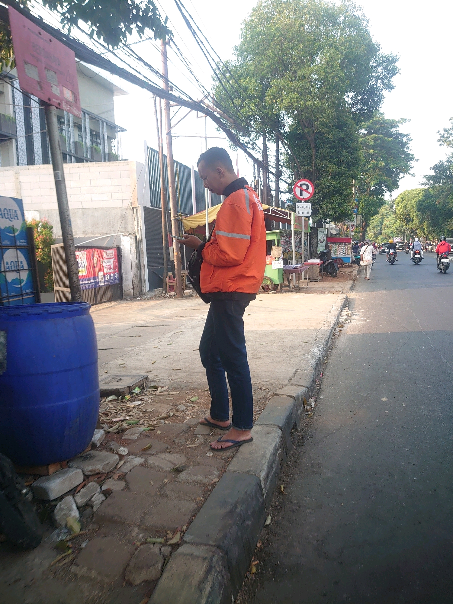 The image captures a street scene in South Jakarta, Indonesia, under clear, bright daylight conditions. A man, likely an online delivery service worker due to his orange jacket with reflective strips, stands on the sidewalk looking at his phone. He is wearing dark pants and flip-flops. To his left, a large blue plastic barrel sits on the edge of the sidewalk, partially obscuring a stacked display of "AQUA" bottled water. Behind the barrel, a building with white brick walls and a facade of glass windows and balconies is visible. Further down the street, a "no parking" sign is posted. A busy road runs alongside the sidewalk, with several motorcycles and a few cars visible in the distance. The street environment appears somewhat urban, with overhead power lines and mature trees lining the road. The sidewalk is paved with concrete and has visible cracks and debris.