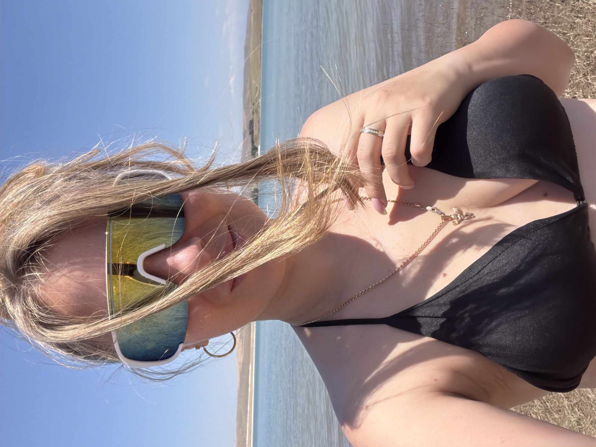 A blonde woman wearing a black bikini and oversized reflective sunglasses stands outdoors in front of a large body of water. The background shows a hilly landscape under a clear blue sky. The woman's hair is windswept, and she is holding her hand up to her chest, revealing a ring on her finger and a gold necklace with a pendant. The image appears to be a selfie, taken from a low angle. The woman is positioned to the left of the frame, with the water and landscape occupying the majority of the background. The lighting suggests it is a bright, sunny day. The location is not discernible from the image itself and cannot be linked to the city of Shymkent, Kazakhstan.