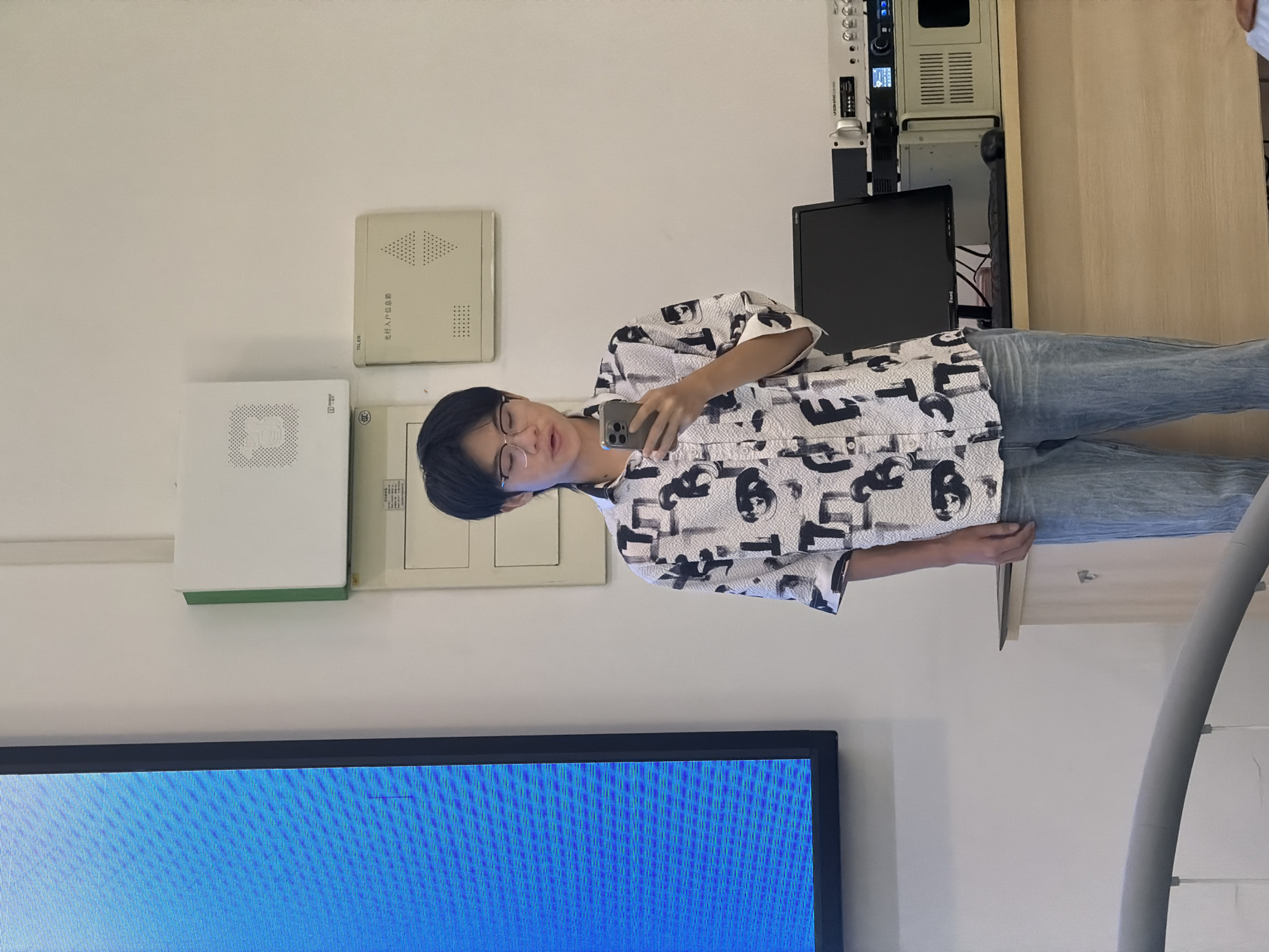 A young East Asian person with dark hair and glasses stands in front of a white wall, holding a smartphone in their right hand and taking a selfie. They are wearing a short-sleeved, collared shirt with a black and white abstract pattern and light blue denim jeans. The background features several wall-mounted items, including two white rectangular boxes with perforated patterns, one with a green border, and a beige rectangular panel with Chinese text. To the person's left is a large, dark-framed screen displaying a blue grid pattern. To their right, on a wooden desk, is a black computer monitor, a keyboard, a mouse, and several pieces of electronic equipment. The image appears to have been taken indoors, likely in an office or classroom setting, possibly in Jiangjin, China.