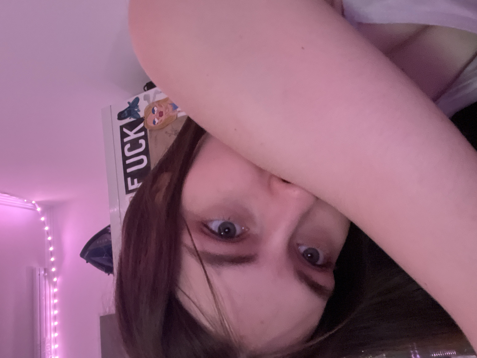A close-up, upside-down selfie of a young person with light skin, brown hair, and blue eyes. The person's right arm is raised, partially obscuring their face. The background features a wall with string lights, a white shelf with stickers (including one with the word "FUCK" on it and a cartoon character), and a dark object mounted on the wall. Outside the window, there are lights, and it could possibly be Saint Petersburg, Russia.