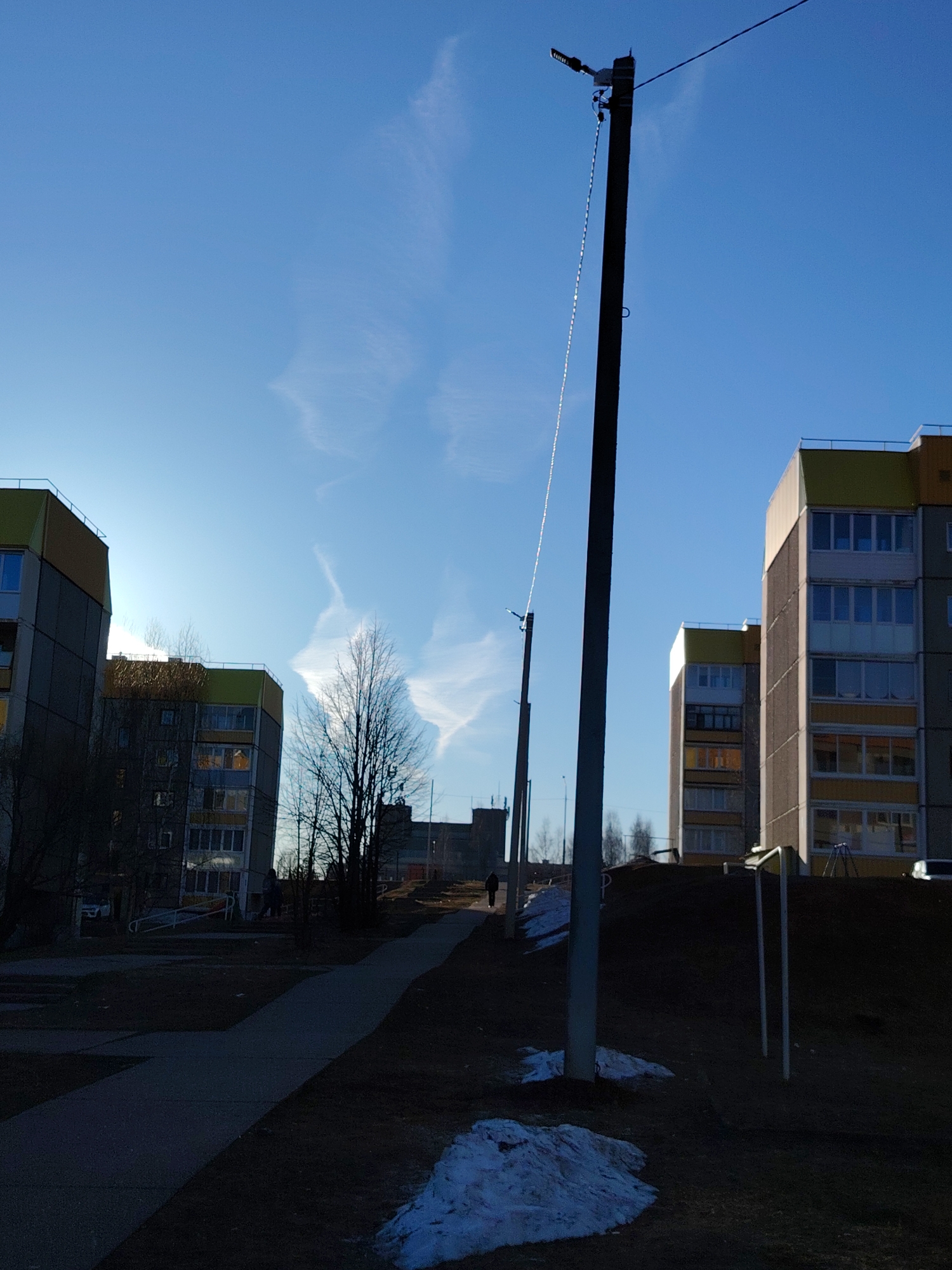 The image depicts an urban scene in Марковский, Russia. The sky is blue with wispy clouds. Several multi-story apartment buildings are visible, with yellow and grey facades. In the foreground, there is a street with a sidewalk, and patches of melting snow on the ground. Several utility poles stand along the side of the street, with a person walking in the distance on the sidewalk. The scene is bathed in sunlight, but large areas are in shadow. Bare trees are visible between the buildings.