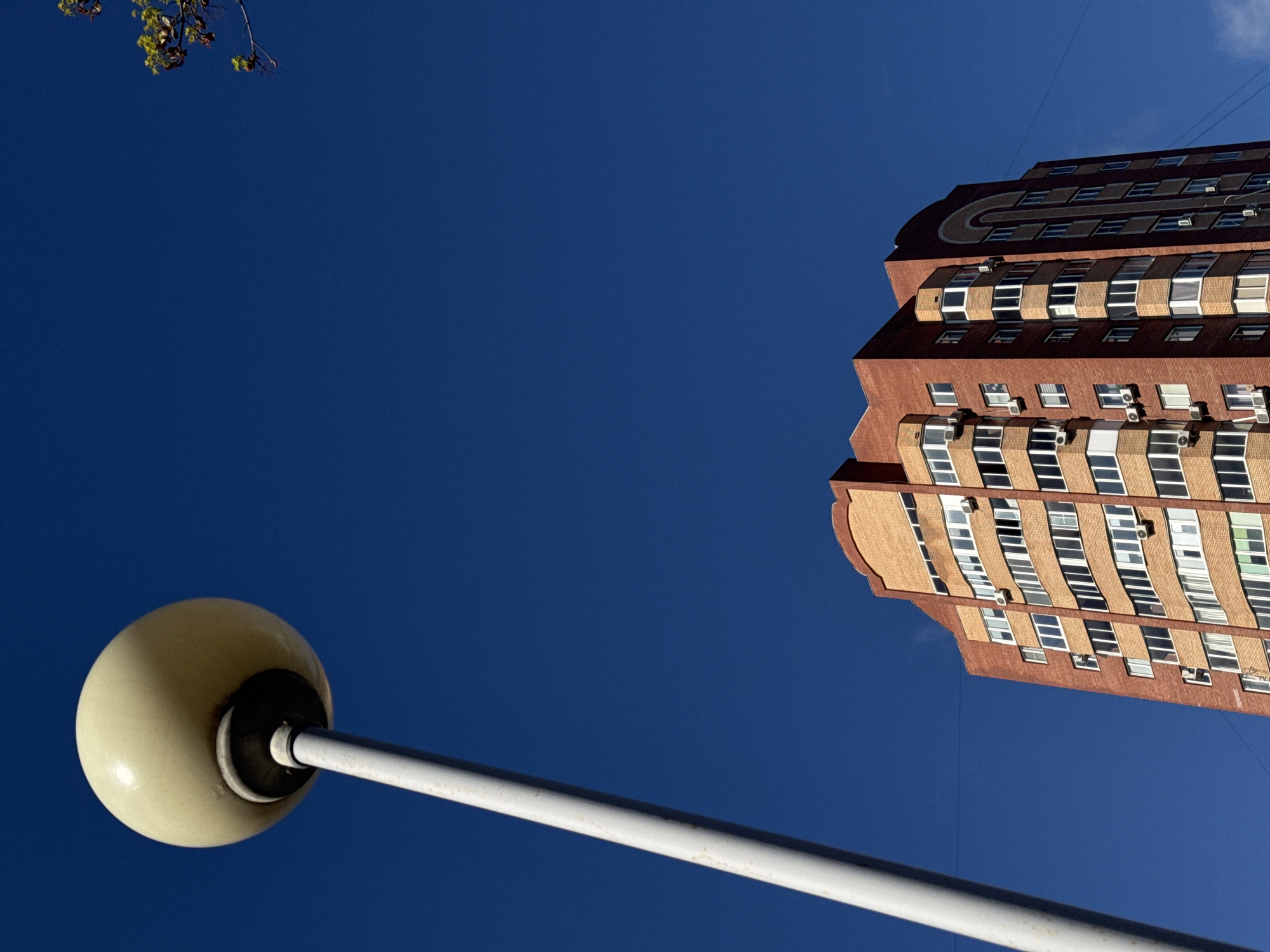 The image presents a striking upward-looking view against a vibrant, clear blue sky, dominated by architectural elements and a street light.
In the upper right portion of the frame, a tall, reddish-brown building with multiple stories rises diagonally. Its facade is characterized by numerous rectangular windows and patterned brickwork in varying shades of brown and beige, creating a textured appearance. Several white air conditioning units are visible attached to the building's exterior. The building's design features receding levels and curved sections, giving it a distinct, almost terraced or stepped profile. Faint lines, possibly power lines or wires, crisscross the blue sky near the building.
A large, spherical, off-white street light occupies the lower left corner, extending diagonally upwards towards the center of the frame. Its white pole is thick and robust, connecting to the globe, which shows some signs of wear or discoloration near its base.
In the top left corner, a small cluster of green leaves on a thin branch is visible, partially silhouetted against the bright sky. The sky itself is a deep, uniform azure, with only a hint of white cloud or intense brightness in the very top right corner.
The perspective is intentionally skewed, capturing the building and street light from a low angle, making them appear to lean into the expansive sky. This unusual composition highlights the geometry and scale of the urban environment. The scene depicts a bright, sunny daytime, with no discernible human activity or visible text.