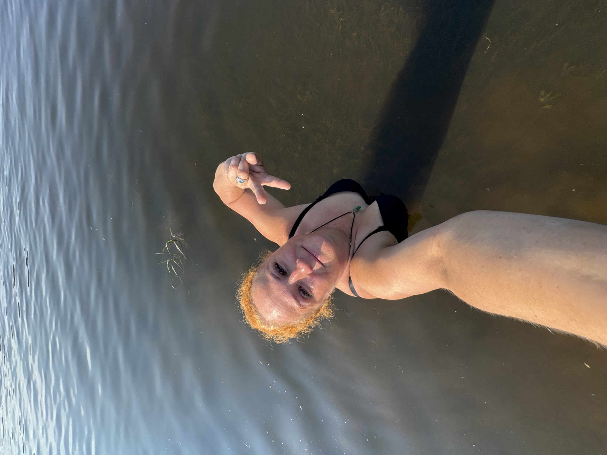 An individual with orange hair, wearing a black bikini, is submerged in dark, murky water, possibly a river or lake. The person is holding the camera in a selfie pose, smiling and looking towards it. Their left arm is extended, and their hand is positioned in a gesture. A ring with a blue stone is visible on their left ring finger. A necklace with a green pendant is worn around their neck. The water is somewhat reflective, showing ripples and a hazy, out-of-focus background. There are some patches of aquatic vegetation visible beneath the surface. Based on the provided context, this scene is located in the city of Chaikovsky, Russia.