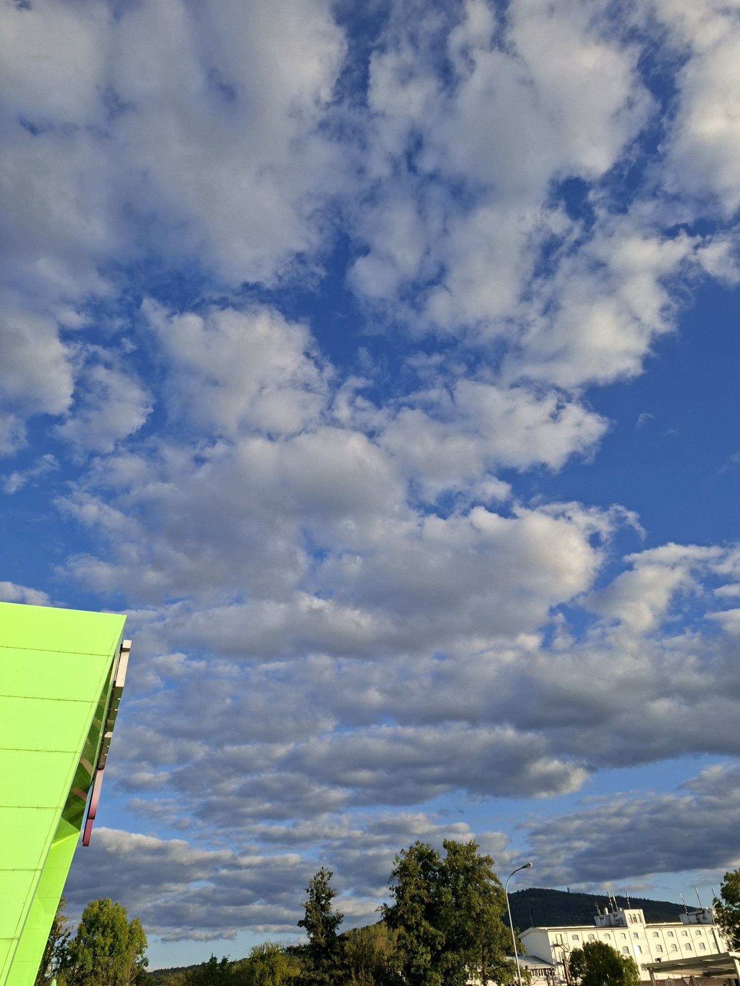 The image captures a bright daytime scene dominated by a vast, dynamic sky filled with fluffy white and grey clouds against a vibrant blue backdrop. In the foreground on the left, a portion of a modern building with a striking lime green, angular facade is visible. The building's surface appears smooth and reflective. Further into the scene, a cluster of lush green trees stands, with one taller, darker evergreen tree positioned to the right of the center. Beyond the trees, a multi-story white building with numerous arched windows and a distinct architectural style, possibly industrial or institutional, is prominent. A streetlamp with a single light fixture rises near the white building. In the distance, a tree-covered hill forms a backdrop, with what appears to be a small tower or antenna visible at its summit. The overall impression is a partly cloudy day in an area that blends modern architecture with natural elements and distant industrial structures.