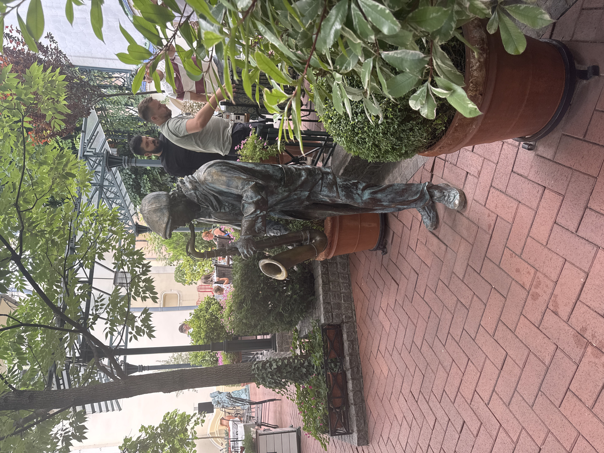 A bronze statue of a saxophonist stands on a red brick patio in Subotica, Serbia. The statue is detailed, showing the musician in a cap and coat, holding a saxophone. Behind the statue, a group of people are seated at tables in an outdoor restaurant or cafe. Trees and bushes provide shade and greenery. The patio is paved with red bricks laid in a herringbone pattern. The environment suggests a pleasant day for outdoor dining.