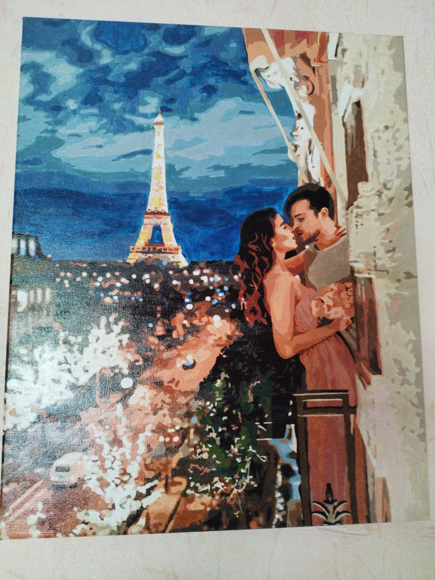 A paint-by-numbers depiction of a romantic scene in Paris, featuring the Eiffel Tower and a couple embracing on a balcony. The night sky is a deep blue with swirling clouds. Below, the city lights are a blur of white and yellow against darker buildings. In the foreground, a man and woman, both with dark hair, are locked in a kiss. The woman wears a pink dress and holds a bouquet of flowers. They are on a balcony with architectural details, including a railing and a column. The painting style is characterized by broad, distinct strokes of color, typical of paint-by-numbers art. The overall impression is one of intimate romance set against the iconic backdrop of Paris at night.