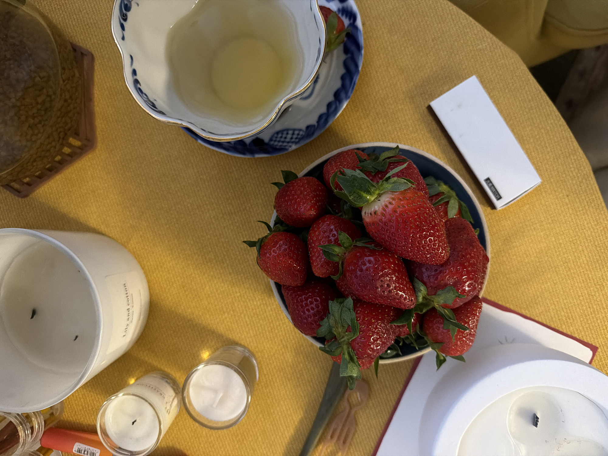 The scene is a tabletop still life with several objects arranged on a mustard-colored surface, possibly a tablecloth or placemat. A bowl filled with fresh strawberries is centrally positioned. To the upper left, there's a teacup and saucer with a blue and white pattern. A small white rectangular box is located to the right of the bowl. Further to the left are several candles, including a large "Lily and Cotton" scented candle and two smaller, lit candles in glass holders. The edges of a red book or notepad can be seen at the bottom right corner. There are no people visible in the scene. The image does not provide any information regarding the location of the scene, especially Moscow, Russia.