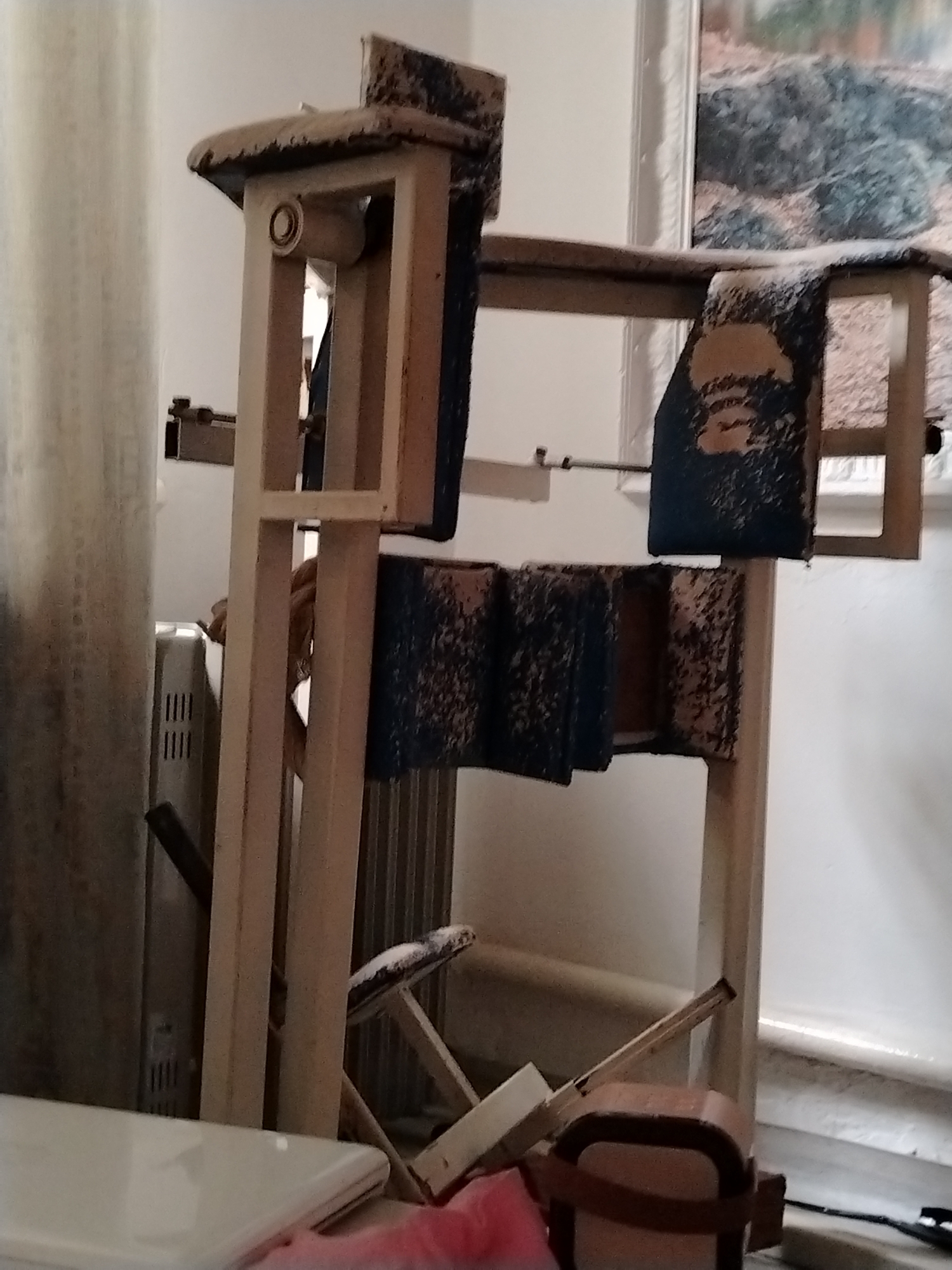 An indoor shot shows a piece of vintage equipment with wooden framing and upholstered pads. The padding appears to be dark blue with a worn, speckled pattern. The wooden frame is light-colored and shows some signs of age and use. The equipment is standing upright in what appears to be a domestic setting, possibly a living room or workshop. Behind the equipment, a radiator is visible, and to the left, a sheer curtain hangs. In the background, a framed picture hangs on the wall, depicting a landscape scene. The lighting suggests an indoor environment with ambient light. There are no people present in the image. The overall scene depicts a well-used, possibly antique, piece of functional equipment in a home environment.