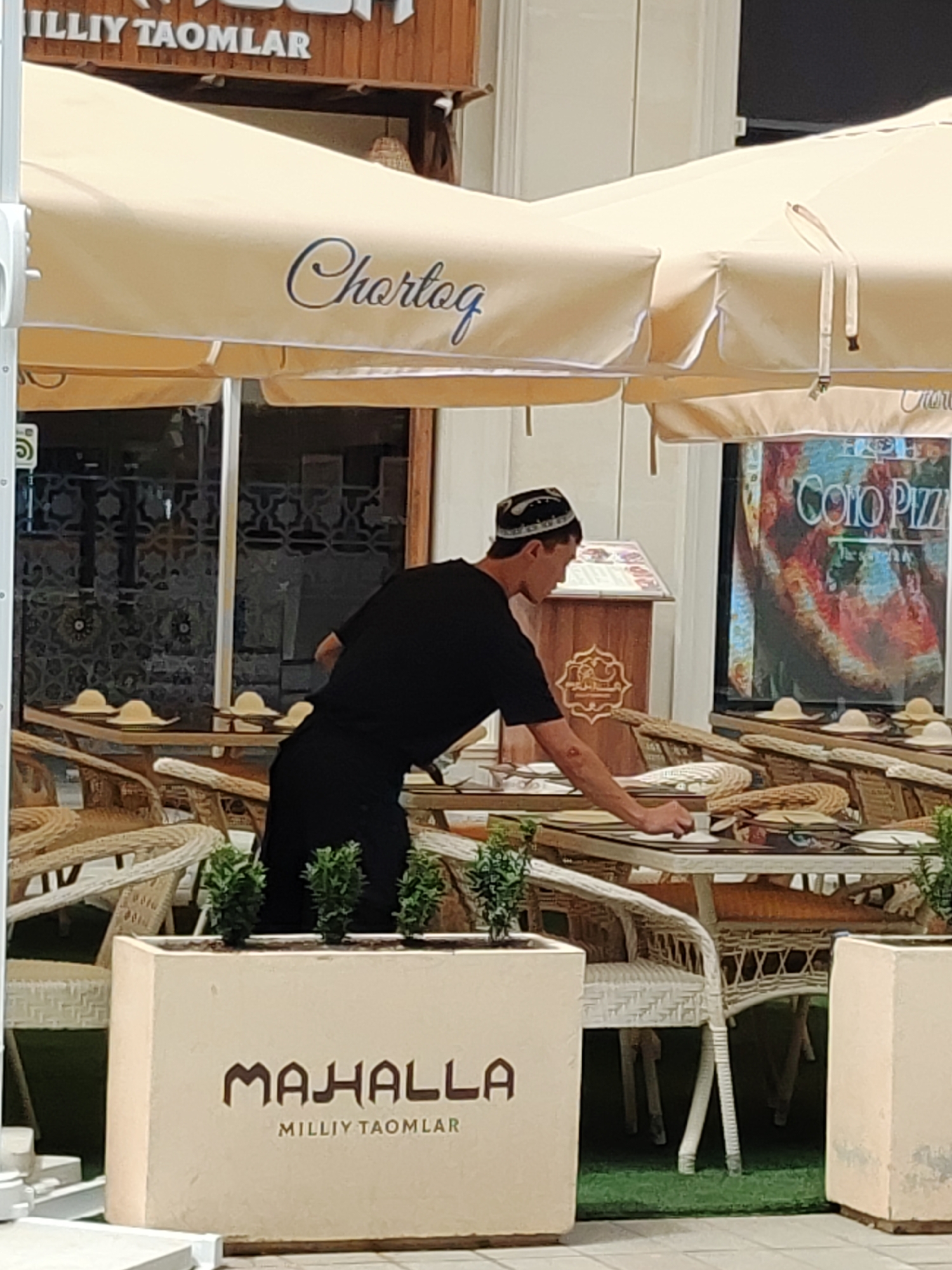 The image shows an outdoor seating area of a restaurant in Tashkent, Uzbekistan. A man wearing a black t-shirt and a skullcap is seen leaning over a table, appearing to be a server or staff member. He is reaching for something on the table, possibly arranging place settings or clearing dishes. The seating area is furnished with wicker chairs and tables, some of which have straw hats on them. In the background, there is a large umbrella with the word "Chortoq" written on it. A sign displaying "MILLIY TAOMLAR" is visible at the top of the building. Another sign with the word "COHO PIZZA" is displayed in a window. A planter box with the word "MAHALLA" and "MILLIY TAOMLAR" inscribed on it is in the foreground. The overall scene suggests a bustling, possibly lunch or dinner service, at a restaurant in Tashkent.