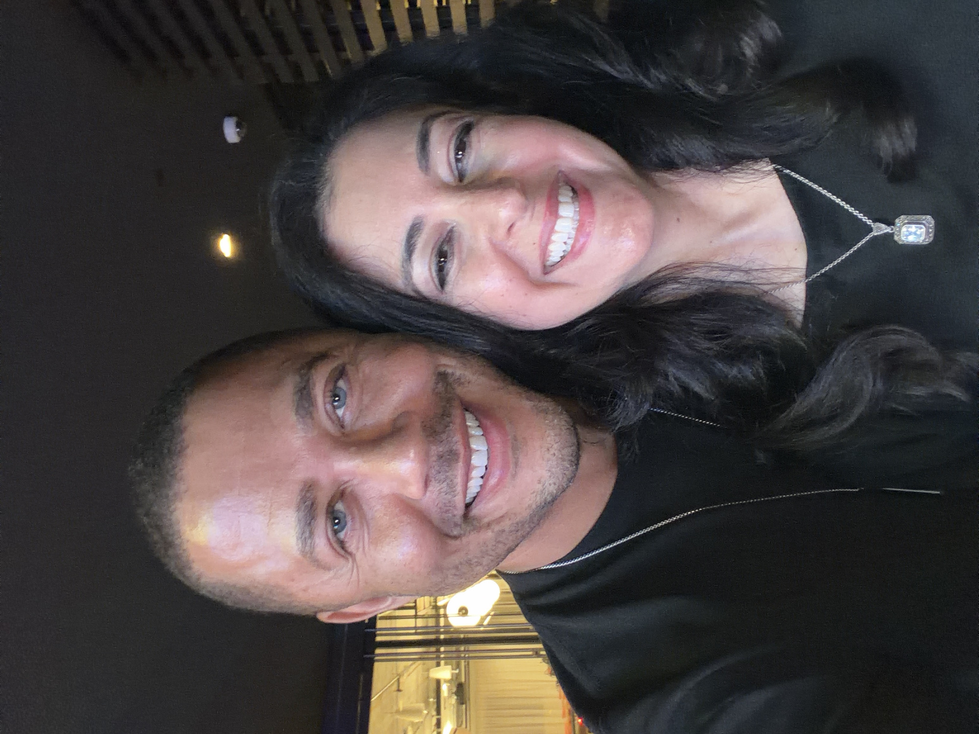 A selfie shows a man and a woman. The man is bald, has blue eyes, and is smiling. The woman has long dark hair and is also smiling. The woman is wearing a silver necklace with a rectangular pendant. The background is dark, with some lighting visible. The location appears to be indoors, possibly a restaurant or bar in an urban environment. The city of Unknown, Unknown cannot be confirmed from the image.