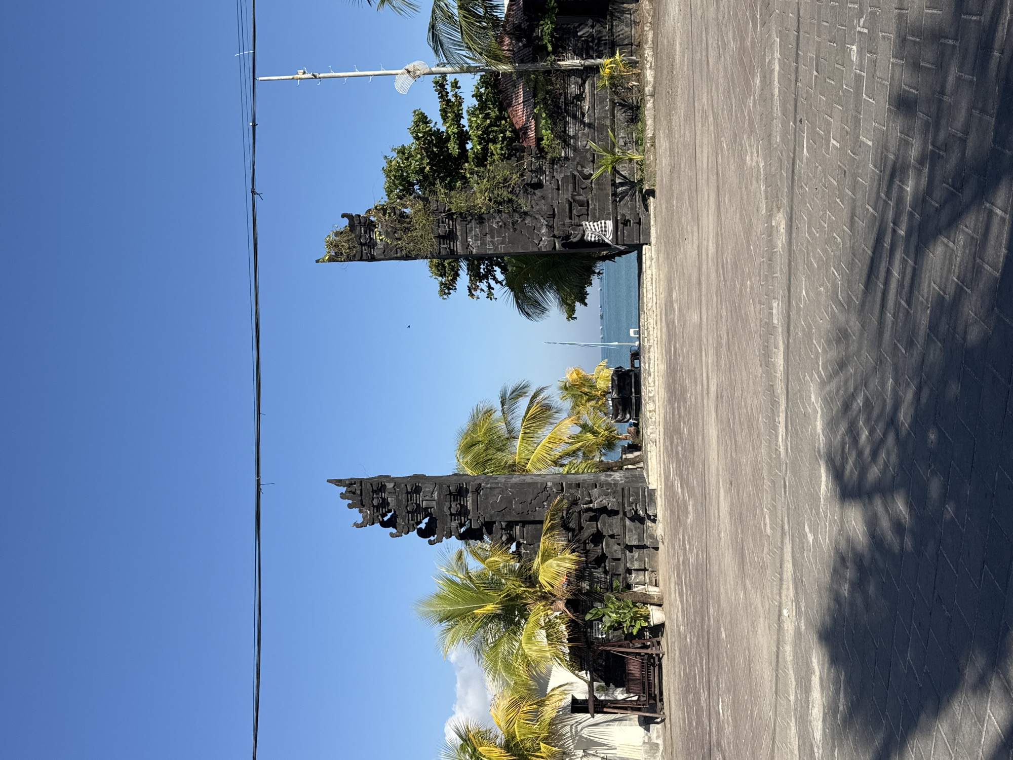 The image depicts the entrance to a beachfront property in Kuta, Indonesia, characterized by a traditional Balinese split gate made of dark stone. The gate stands prominently in the center of the frame, flanked by lush palm trees with vibrant green fronds. Beyond the gate, a calm turquoise sea stretches towards the horizon, dotted with a few boats. The foreground features a paved area, possibly a parking lot or courtyard, with a distinct shadow cast by palm leaves. To the left of the gate, a wooden swing set is visible, adding a touch of leisure to the scene. The sky is a clear, brilliant blue, indicating a sunny day. The overall atmosphere is serene and tropical, characteristic of a seaside resort.