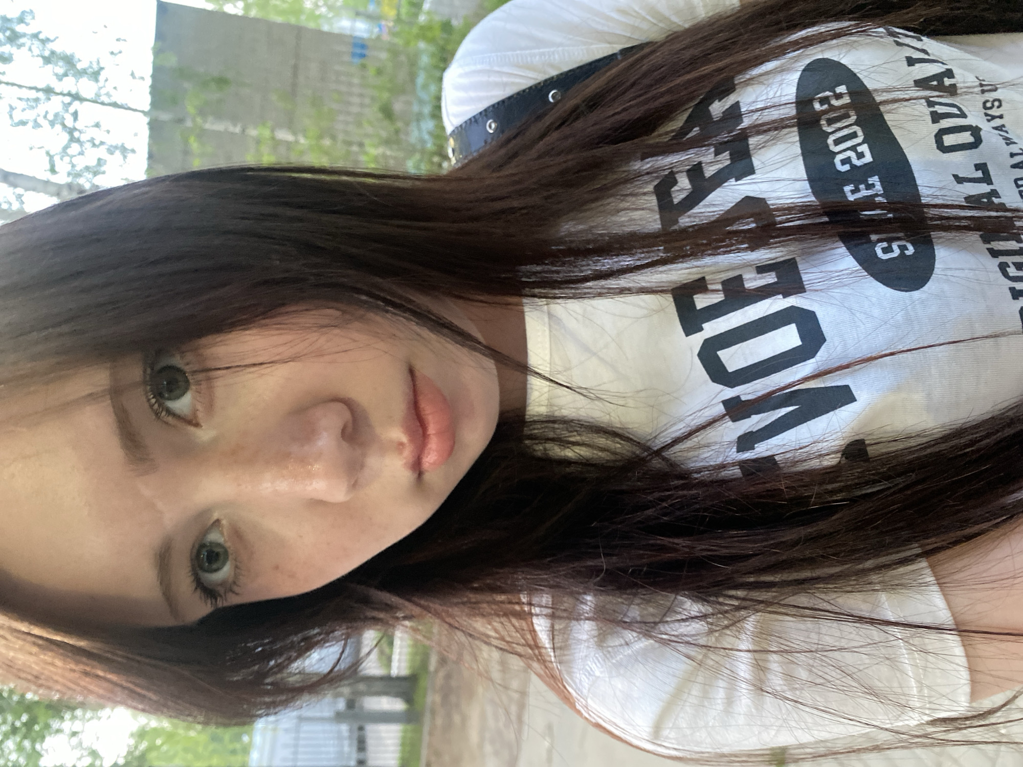 A close-up, vertically oriented photograph shows a young woman with light skin and dark brown hair, wearing a white t-shirt with black lettering, seemingly outdoors. The lettering on the shirt reads "VOICE OF...SINCE 2002...RIGINAL QUALITY...ALWAYS UNIQUE." The woman has freckles on her nose and green eyes. In the background, there is greenery and a building. The location is undetermined.