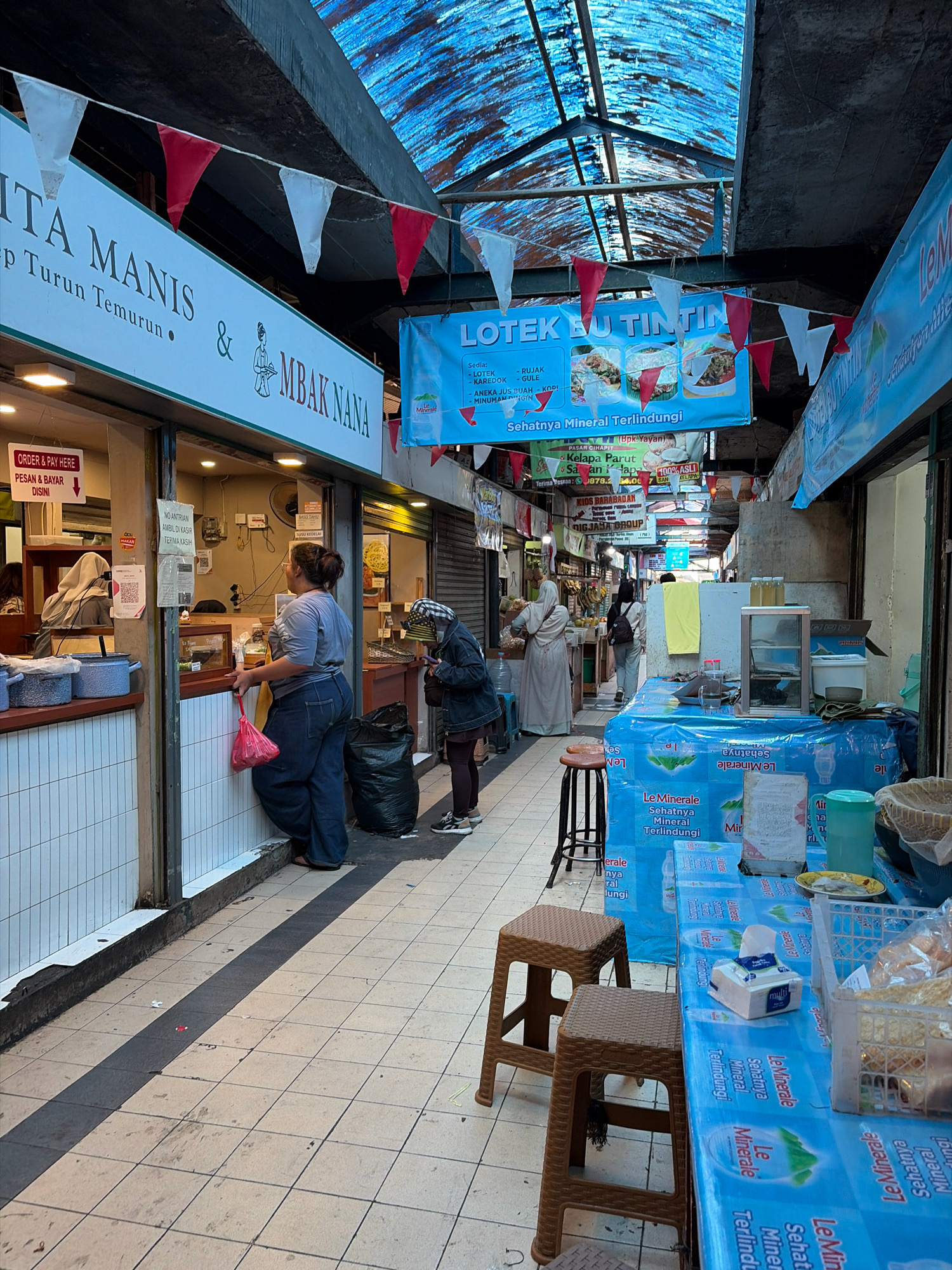 The image depicts a market scene in Bandung City, Indonesia, likely a food stall area with a covered walkway. In the foreground, two brown plastic stools sit in front of a table draped with a blue cloth displaying branding for "Le Minerale" bottled water. Various items are on the table, including a tissue box, plastic containers, and a woven basket. To the left, a woman in a gray t-shirt and blue jeans stands at a counter, holding a red plastic bag, appearing to interact with someone behind the counter. A black garbage bag is visible next to her. Further down the walkway, other individuals are seen, some walking and others browsing at stalls. Banners and signs hang above, indicating various food and drink offerings such as "LOTek Bu Tintin." The walkway is covered by a corrugated plastic roof that allows light to filter through, creating a dappled effect on the tiled floor. The overall impression is of a bustling local market.