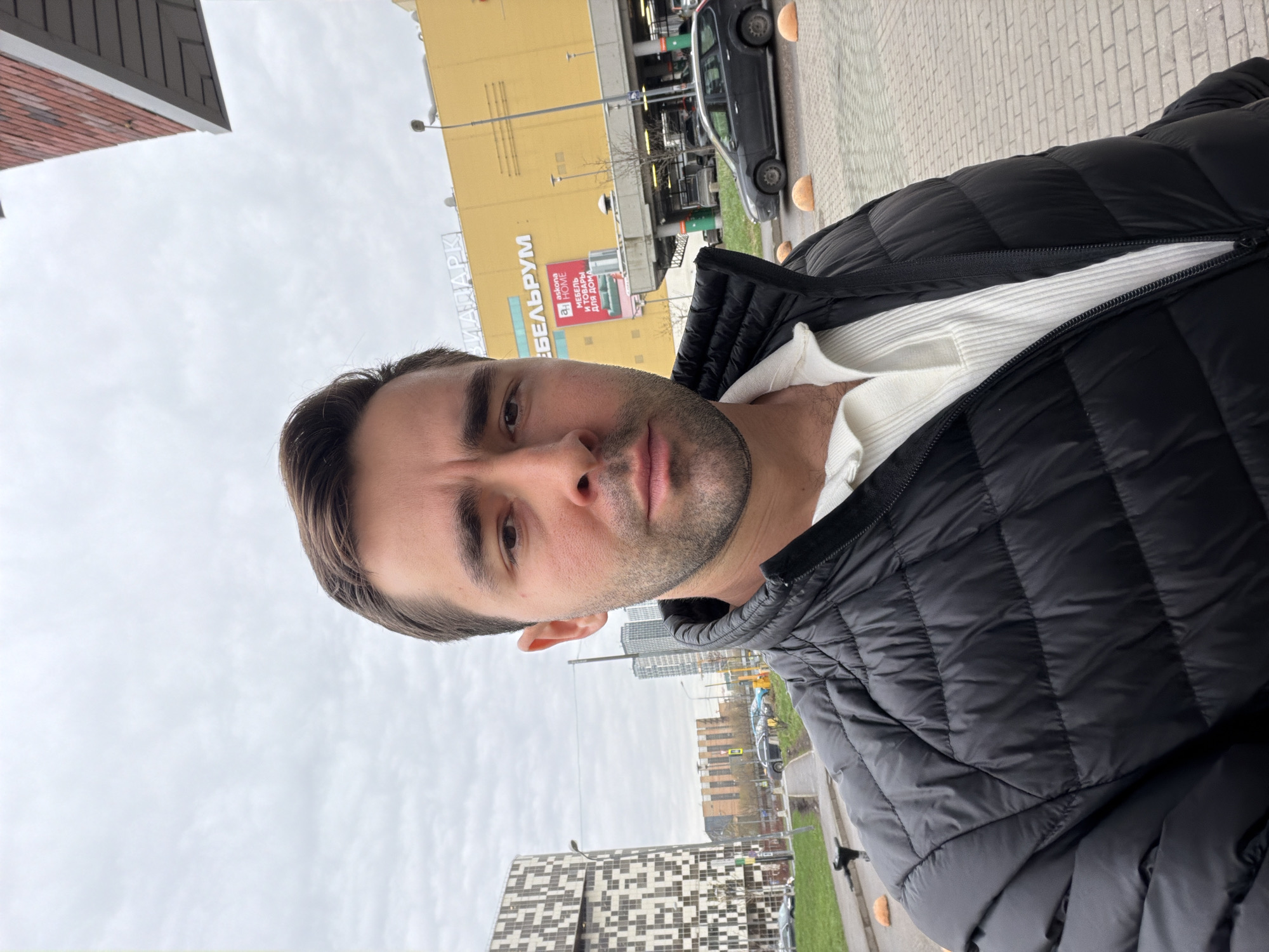 A man is pictured in a selfie against the backdrop of a Moscow city scene. He is wearing a black puffer jacket over a white top. In the background, a building with "БЕЛЬРУМ" written on it is visible, along with cars and other urban structures. The weather appears overcast. Other buildings are visible further away, suggesting an urban environment.