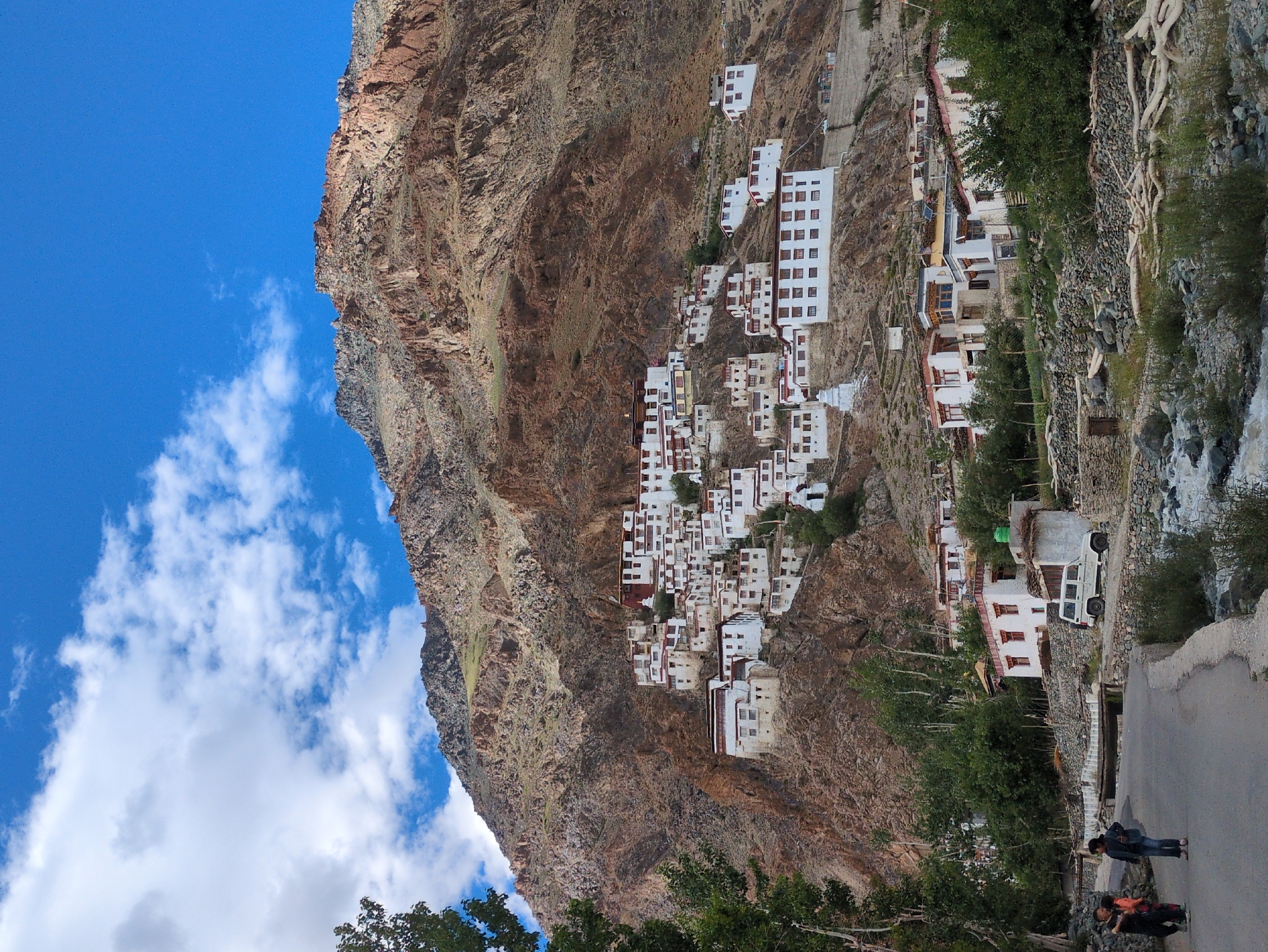 This is a medium-shot, vertical, eye-level, full-color photograph depicting a Tibetan Buddhist monastery nestled on the side of a rocky mountain in Yulang, India. The monastery's white, multi-storied buildings with dark-trimmed windows are spread across the mountainside, appearing as a collection of connected structures. The mountain itself is rugged and barren, with shades of brown and ochre dominating its surface, and sparse green vegetation clinging to its lower slopes. A clear blue sky with scattered white clouds forms the backdrop. In the foreground, a paved road curves through the scene, with three individuals, two adults and a child, walking on it. To the right of the road, a white SUV is parked near a cluster of buildings, and a rocky stream flows beside the road.