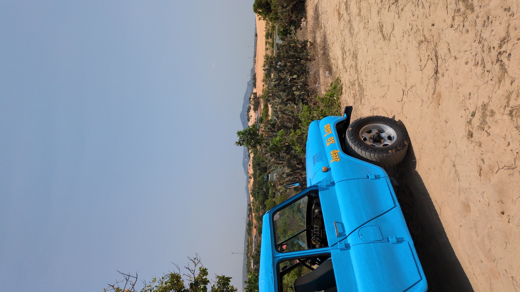 A bright blue vintage Jeep, labeled "Jeep Xo Loan," is parked on a sandy surface, possibly a dune, in Phuoc Dinh, Vietnam. The vehicle is oriented with its right side facing the viewer, showcasing the passenger side door and a tire. The landscape beyond the jeep includes green vegetation, sandy dunes, and a distant skyline. The sky is a clear, pale blue.
