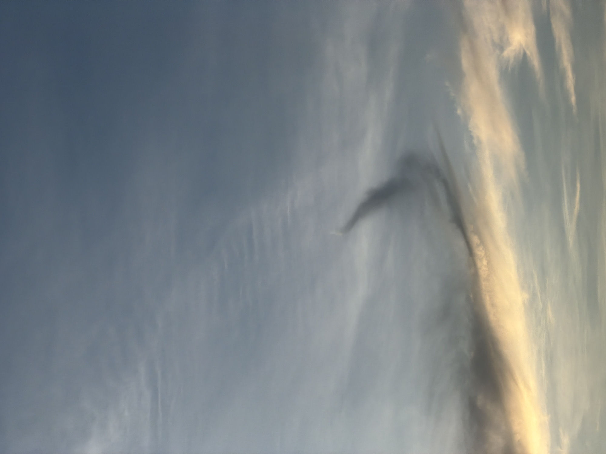 The image shows a sky with wispy clouds. A distinct, dark, comma-shaped cloud formation is visible in the center, curving upwards. The lower portion of the image is illuminated by a warm, golden light, suggesting either sunrise or sunset. This scene is set against the backdrop of the sky above Hoi An, Vietnam. There are no people, subjects, objects, or actions depicted in the image.