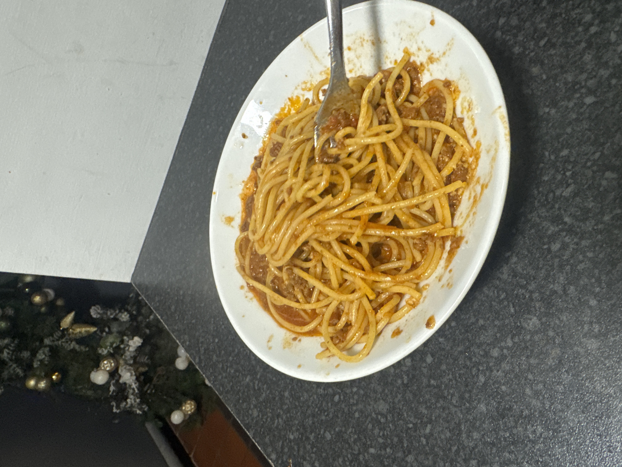A partially eaten plate of spaghetti with meat sauce sits on a dark gray countertop. A silver fork is inserted into the noodles. Part of a Christmas tree decoration is visible in the lower left corner of the image. The countertop is next to a white surface. The location is Warrington, United Kingdom. There are no people visible in the image.