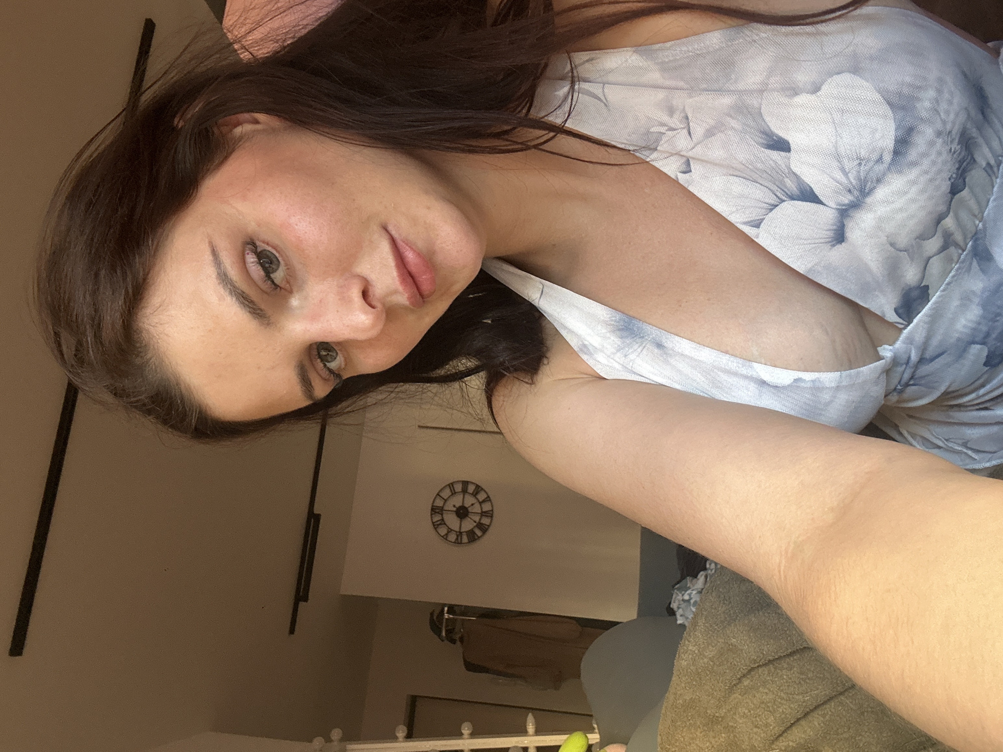The media file is a portrait of a young woman taken indoors. She has long, dark brown hair and is looking directly at the camera with a neutral expression. She is wearing a light-colored top with a floral pattern. The background is dimly lit and appears to be a domestic setting. There is a clock visible on the wall behind her, and the overall lighting suggests it could be evening or a room with artificial light. The image is oriented vertically, with the woman's head on the left side of the frame and her body extending to the right. The focus is primarily on her face and upper body.