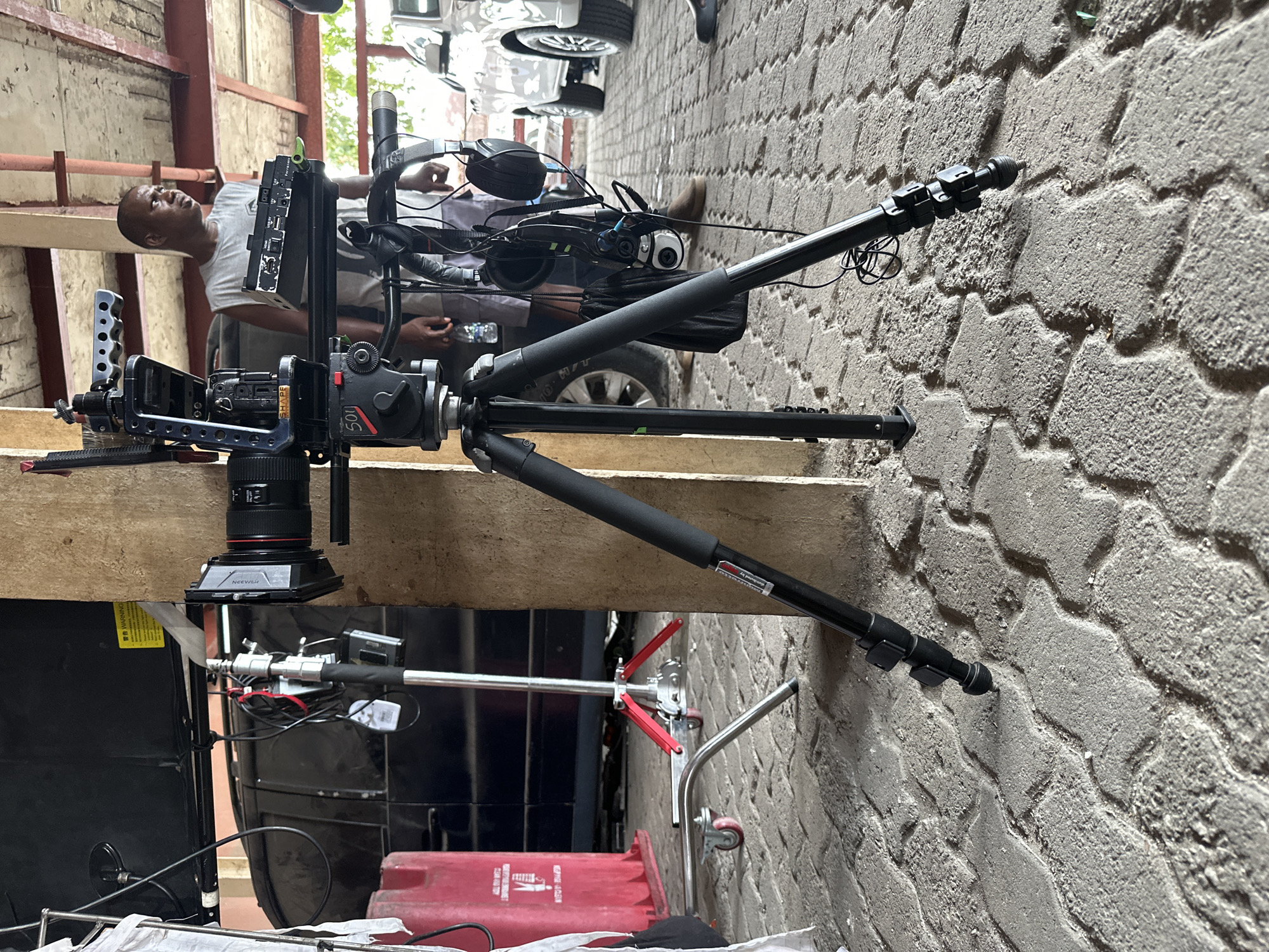 The image shows a film set, possibly located in a constricted urban area, in Lagos, Nigeria. The primary focus is on camera equipment, including a professional video camera mounted on a tripod. The tripod is positioned on a wooden platform. A black male is visible to the left of the camera, presumably working on the set. Other pieces of equipment, including a dolly and what appears to be a generator, are present near a brick wall. A vehicle is visible above the set.