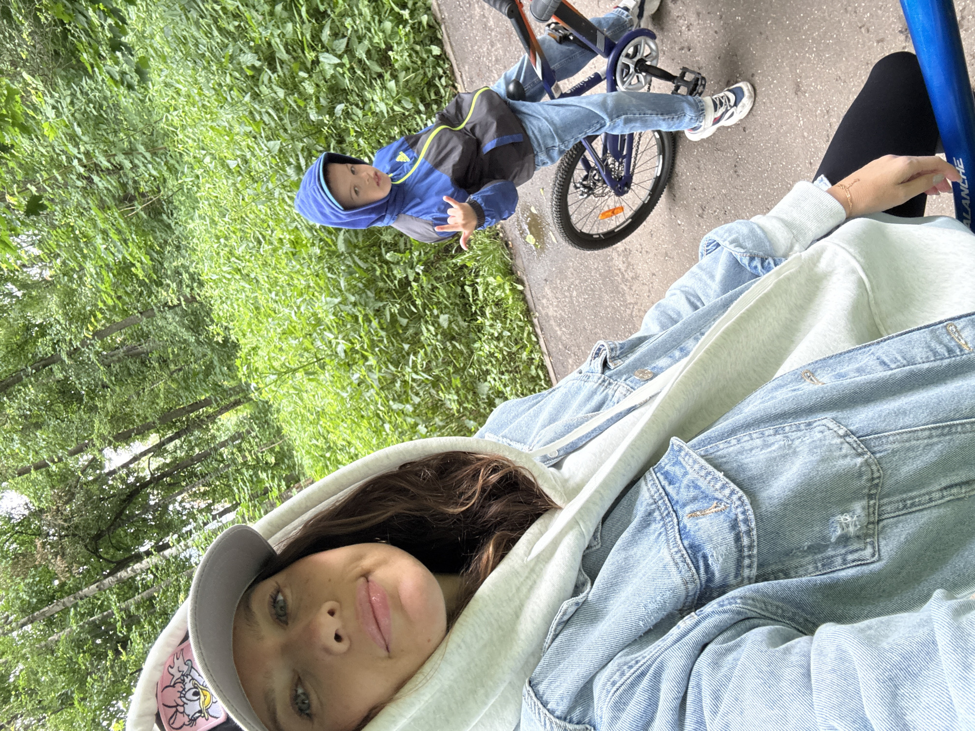 A woman and a young boy are in a park. The woman is in the foreground, taking a selfie. She is wearing a denim jacket and a grey hoodie. The boy is on a bicycle behind her, and he is wearing a blue and black jacket and jeans. The park has trees and greenery. There is a paved path, and it looks like it might be a cloudy or overcast day. The photo seems to have been taken in Moscow, Russia.
