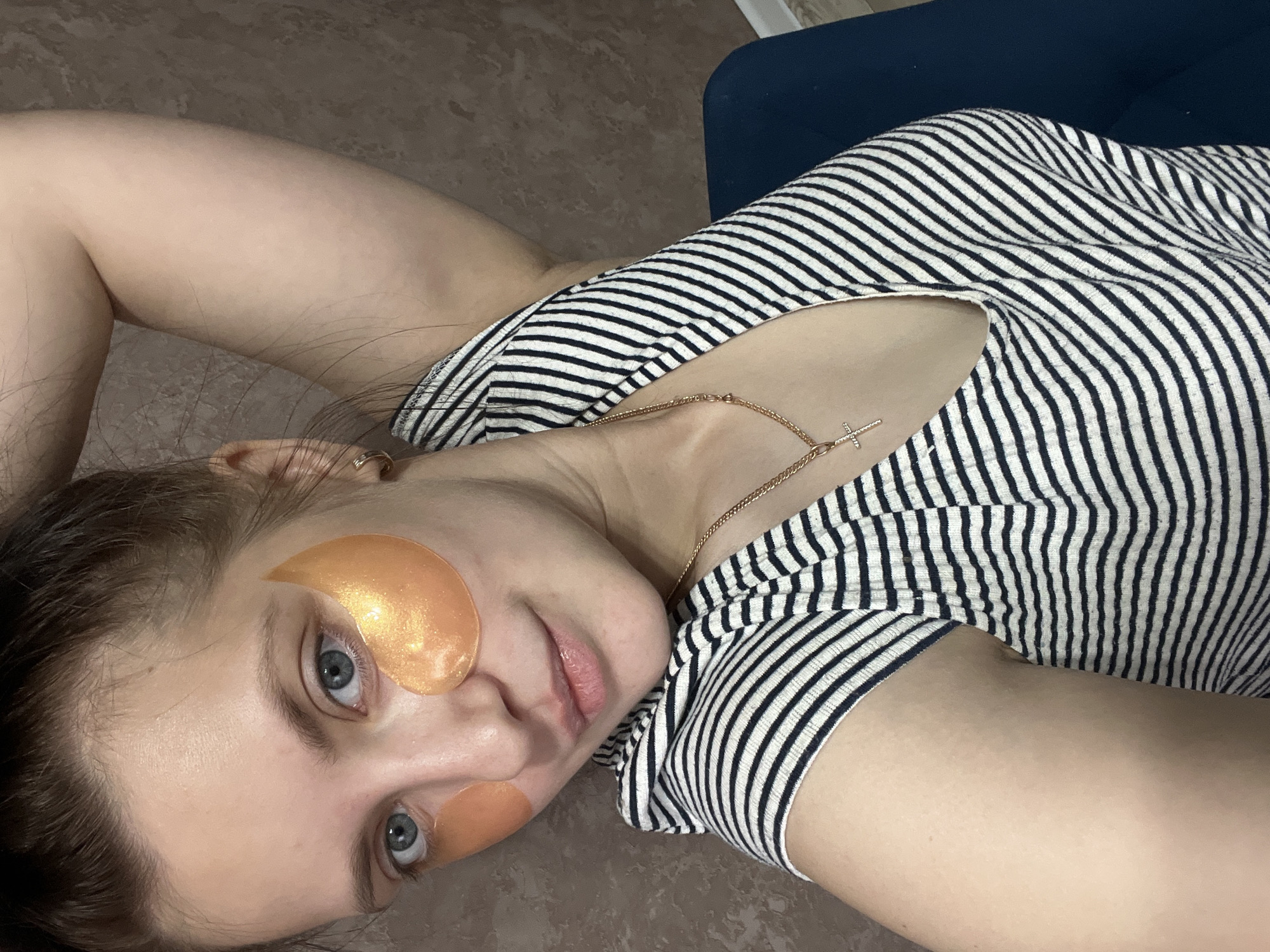 A woman with light skin, blue eyes, and dark hair is lying on a brown, reflective surface, likely a floor. She is wearing a striped black and white shirt and a gold necklace with a cross pendant. She has two gold-colored eye patches under her eyes. Her right arm is bent and positioned behind her head. Part of a dark blue object, possibly furniture, is visible in the upper right corner of the frame. The environment is indoors. The location of the scene is Марковский, Russia.
