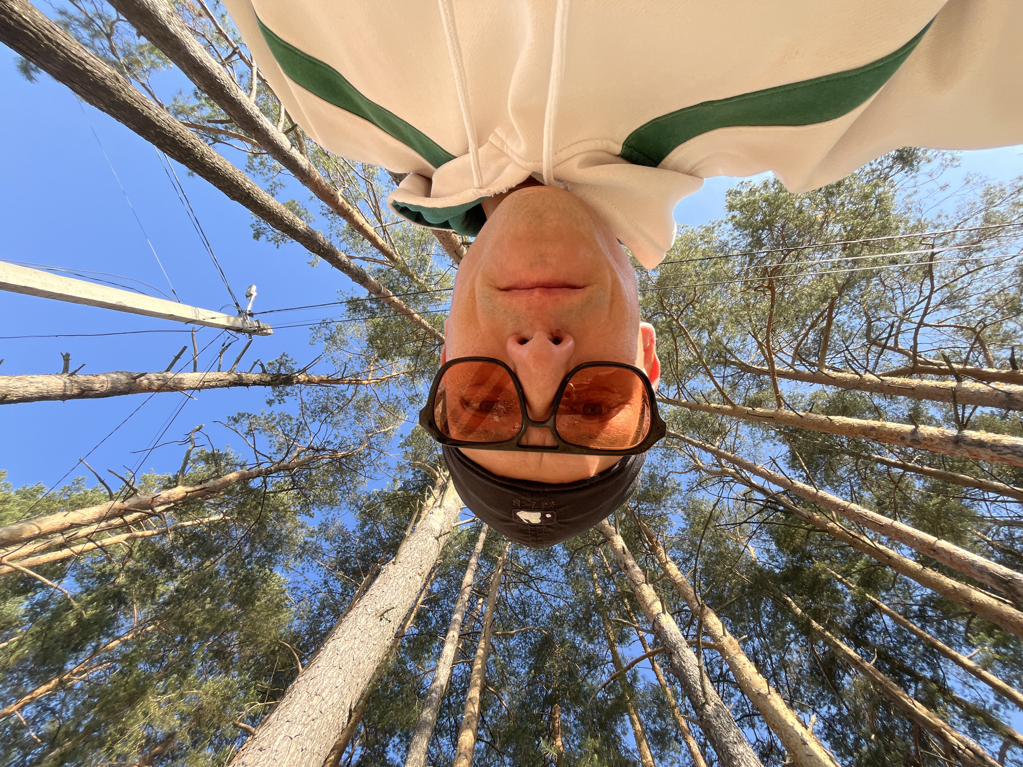 A low-angle photograph shows a person looking upside down at the camera, against a background of tall trees and a bright blue sky. The person is wearing glasses with amber-tinted lenses and a dark-colored baseball cap. Their clothing is a light-colored hoodie with a green trim around the hood. Tall, thin trees rise into the frame. Some powerlines and a utility pole are visible in the background. The photo was taken in Chaikovsky, Russia.
