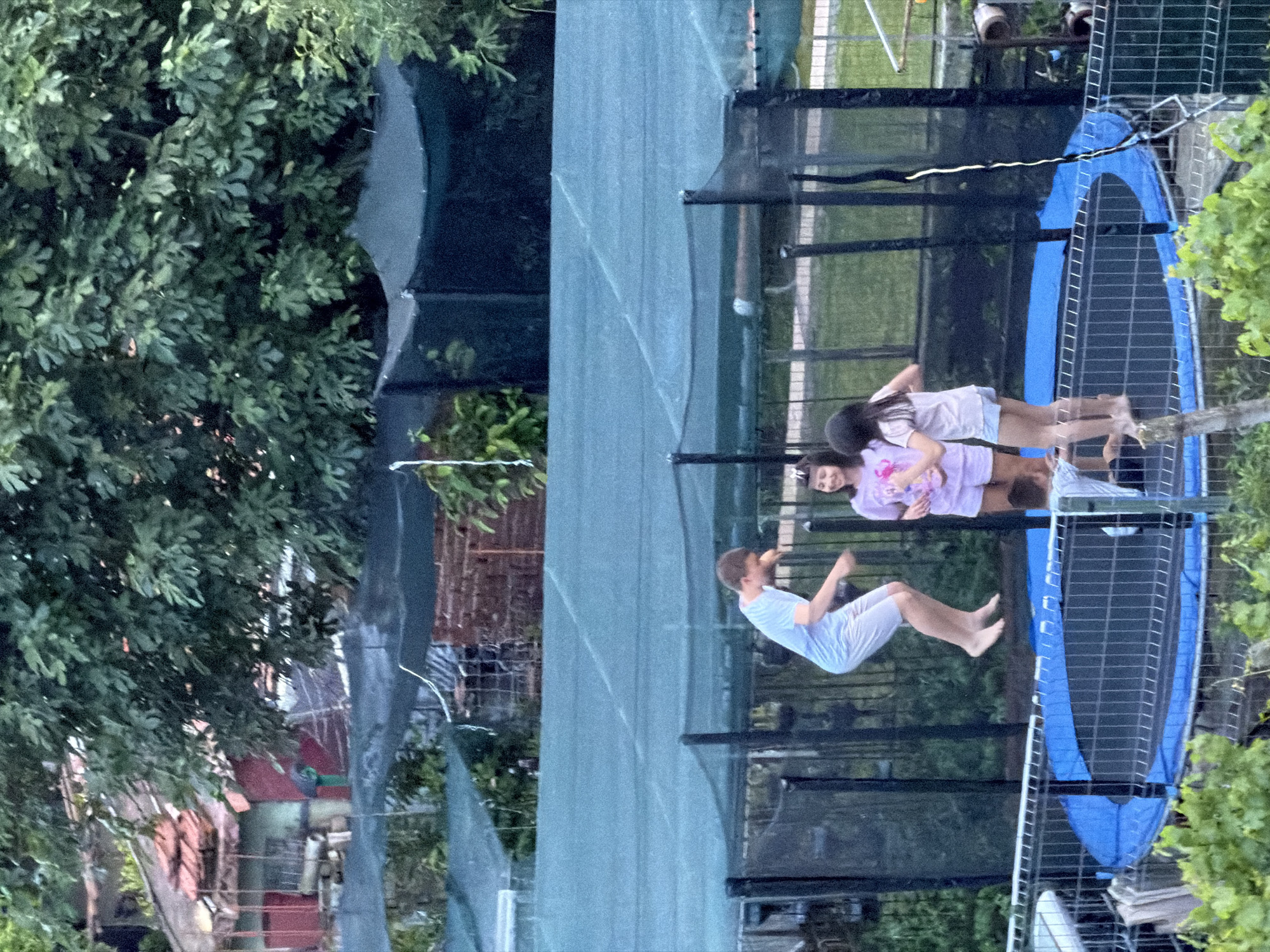 The image depicts four individuals on a trampoline with a blue safety pad and a black net enclosure. Two girls are standing near the edge, while a man is in mid-air, seemingly jumping. A fourth person is visible between the two girls.
The trampoline is situated outdoors, likely in a backyard setting, with lush green foliage surrounding it. A building with a red roof is partially visible in the background. The location is not specified but it is in the city of Burgas, Bulgaria. The weather appears to be overcast and it is daytime.
