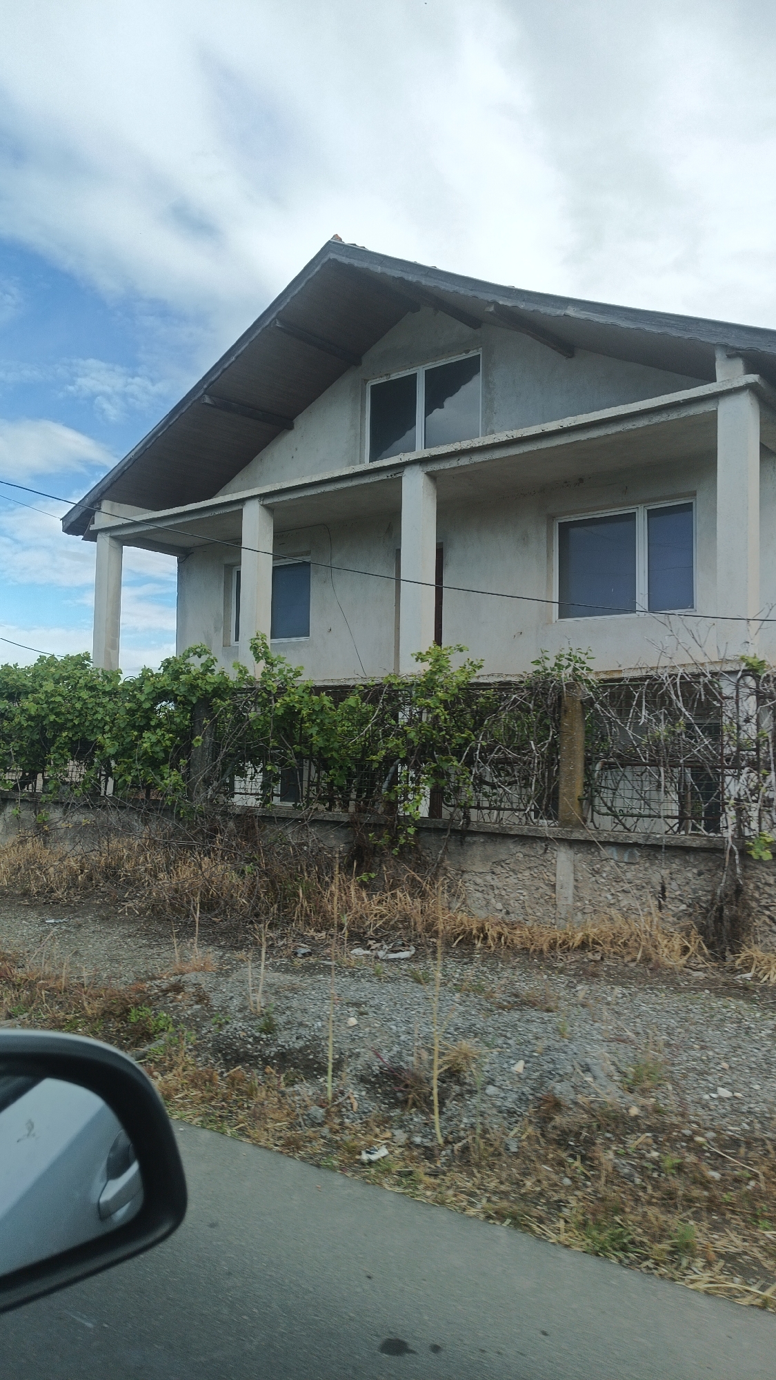 The image depicts a two-story building with a concrete structure and a gabled roof. The building has a second-story balcony supported by columns. Windows are visible on both floors. A fence runs along the property line, partially obscured by overgrown vegetation. The ground in front of the building is covered in gravel and dry vegetation. A portion of a vehicle's side mirror is visible in the lower left corner of the image. The sky above the building is cloudy. Based on the prompt, the location is Milevo, Bulgaria. There are no people visible in the image.