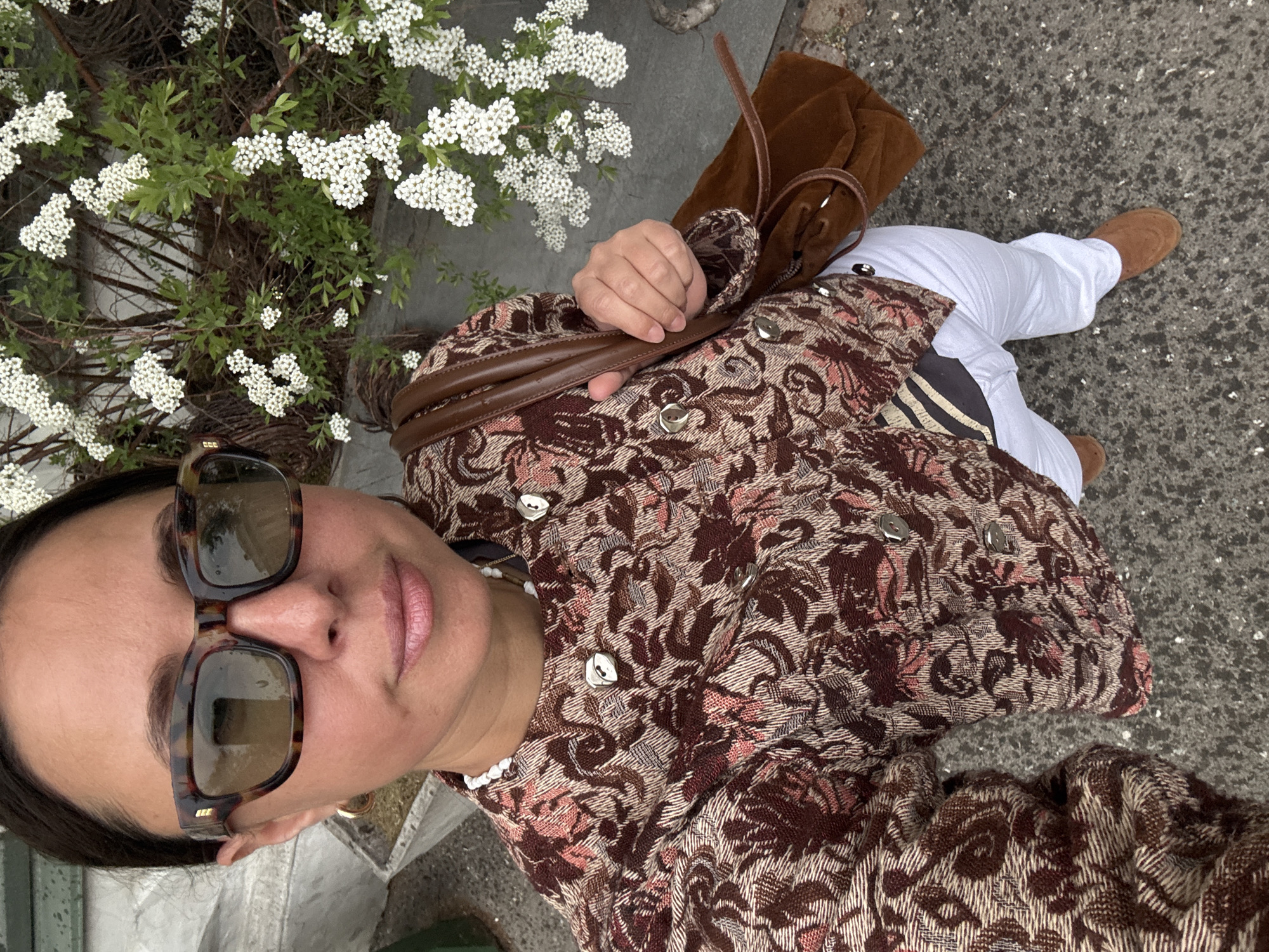 A woman is shown from an overhead perspective, likely taking a selfie. She is wearing sunglasses, a brown floral patterned jacket, a white beaded necklace, white pants, and brown suede shoes. She is holding a brown leather handbag. Beside her is a bush of small white flowers. The background is a concrete surface. There is no clear indication of this image being taken in the city of Kazan, Russia.