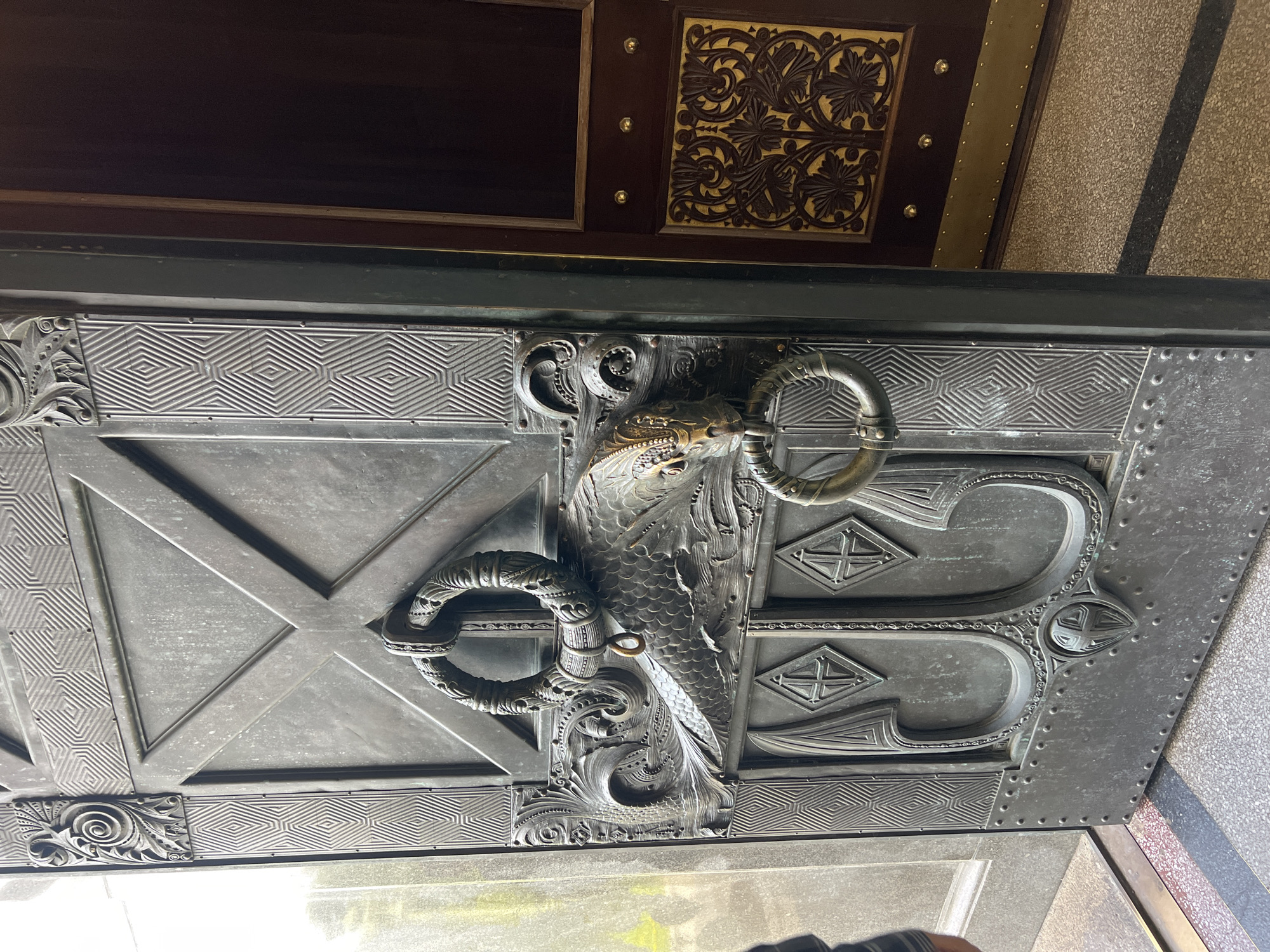 An ornate, dark metal door, likely made of bronze, is ajar, revealing a glimpse of a second, darker wooden door within a building. The prominent metal door is heavily decorated with intricate relief carvings. The center of the door features a large, sculpted dragon or sea serpent design, with its head grasping a ring-shaped handle. Another ring handle is suspended from the serpent's neck. Above and to the sides of the serpent are panels with geometric patterns, composed of diagonal lines forming diamonds. Further decorative elements include ornate scrolling motifs on the left side of the door and circular embellishments with what appear to be cross-like designs at the bottom. The floor in front of the door is tiled with a speckled pattern. The exterior environment visible to the left of the door is blurred and appears to be a brightly lit outdoor space, possibly with some foliage. A person's leg and lower torso, wearing dark pants and a striped shirt, are visible on the far left edge of the frame, partially obscured by the open door. The context of Kronstadt, Russia, suggests this may be an entrance to a historical or governmental building, given the elaborate craftsmanship.