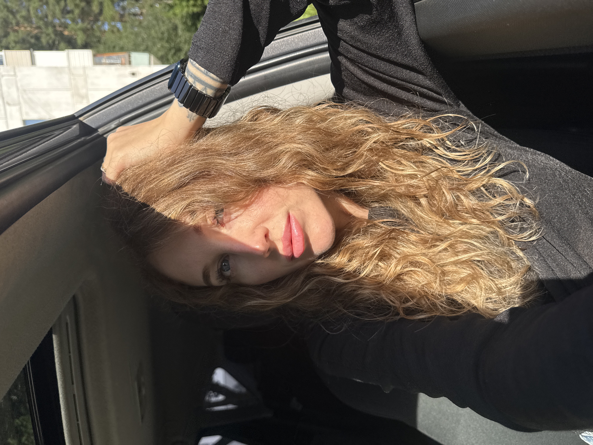 The media file shows a close-up, sideways portrait of a woman with long, wavy blonde hair. She is wearing a black long-sleeved top and a black bracelet on her left wrist. Her left hand rests on her head, and her right arm is bent, with her hand near her face. Sunlight is illuminating her face, creating a strong highlight on her cheek and forehead. She appears to be looking towards the camera with a neutral expression.
The setting appears to be inside a vehicle, suggested by the car door frame visible on the left and the interior car paneling in the upper right. Outside the car, greenery and some white structures are visible, indicating an outdoor environment. The bright sunlight suggests it is daytime, likely late afternoon given the angle of the light. The overall mood is relaxed and candid. There is no visible text in the image.