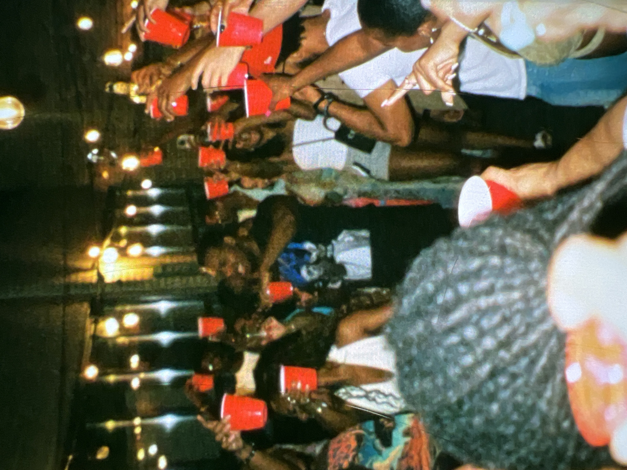 The image captures a lively gathering indoors, likely a party or social event, with a festive atmosphere. A group of people, predominantly young adults, are seen raising red plastic cups, suggesting a toast or celebration. The lighting is dim, illuminated by numerous warm-toned string lights that hang overhead, creating a bokeh effect. In the foreground, a person's head with intricate braided hair is visible, obscuring some of the scene. Several individuals in the middle ground are distinctly seen holding red cups, with some wearing casual attire. One woman in the middle right is wearing a white strapless top and shorts, and carrying a black crossbody bag. The background reveals more individuals and the textured brick walls of the venue. The overall impression is one of camaraderie and enjoyment. The image context implies a vibrant social scene within Lagos, Nigeria, given the cultural context of such gatherings.