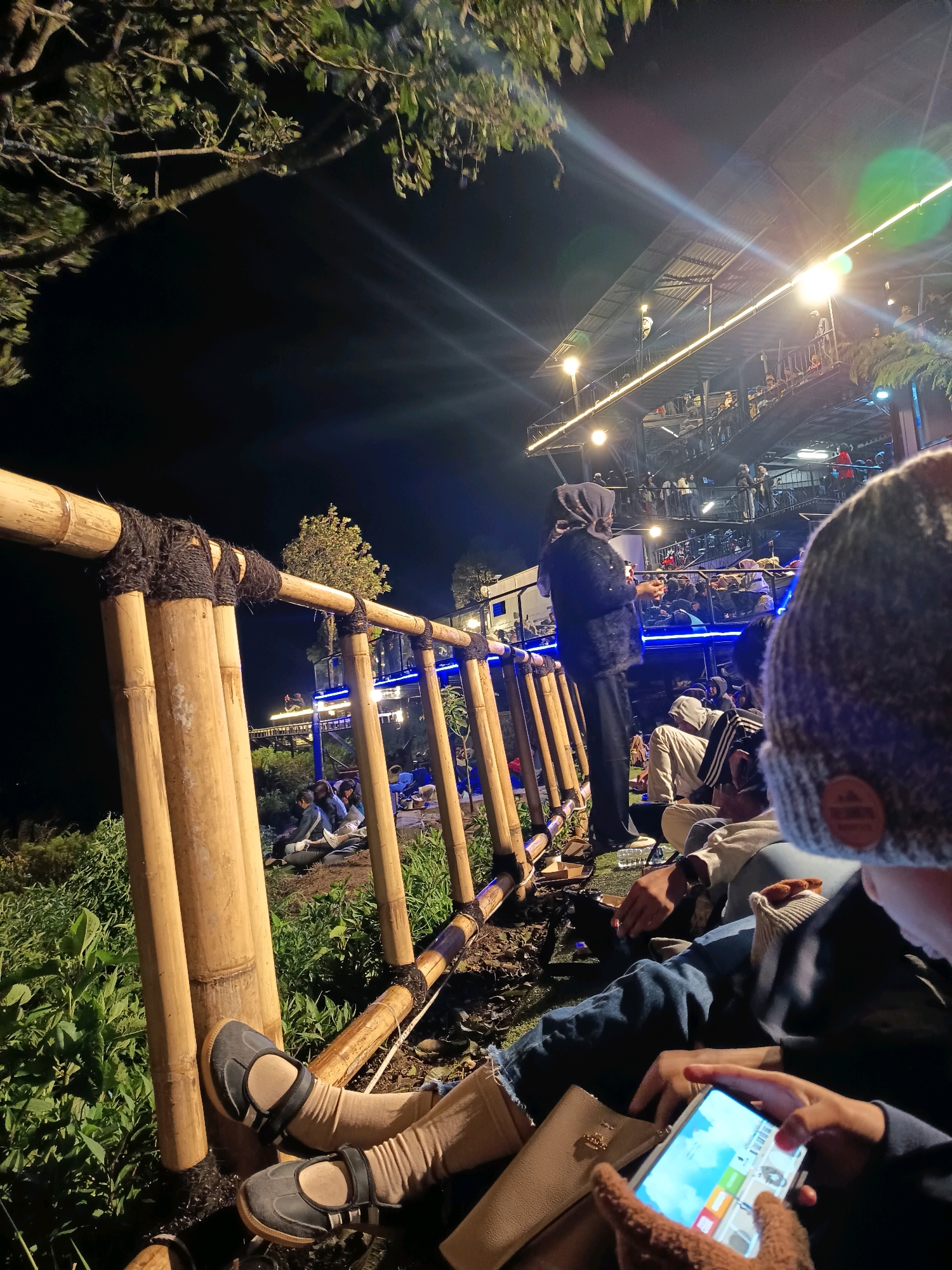 The image depicts an outdoor evening scene at what appears to be a popular gathering spot in Keditan, Indonesia. In the foreground, a person wearing a grey knit hat and a denim jacket is holding a smartphone. Their legs, clad in light brown stockings and dark shoes, are resting near a bamboo railing. Behind them, other individuals are seated or standing, some engaged in conversation. The background reveals a multi-level structure illuminated with warm lights, suggesting a restaurant, cafe, or entertainment venue. Several people are visible on different levels of this structure, and blue lighting accents along a walkway add to the ambiance. Lush green foliage surrounds the area, and the dark night sky indicates it is nighttime. The overall atmosphere is lively and social, with an emphasis on outdoor dining or relaxation.