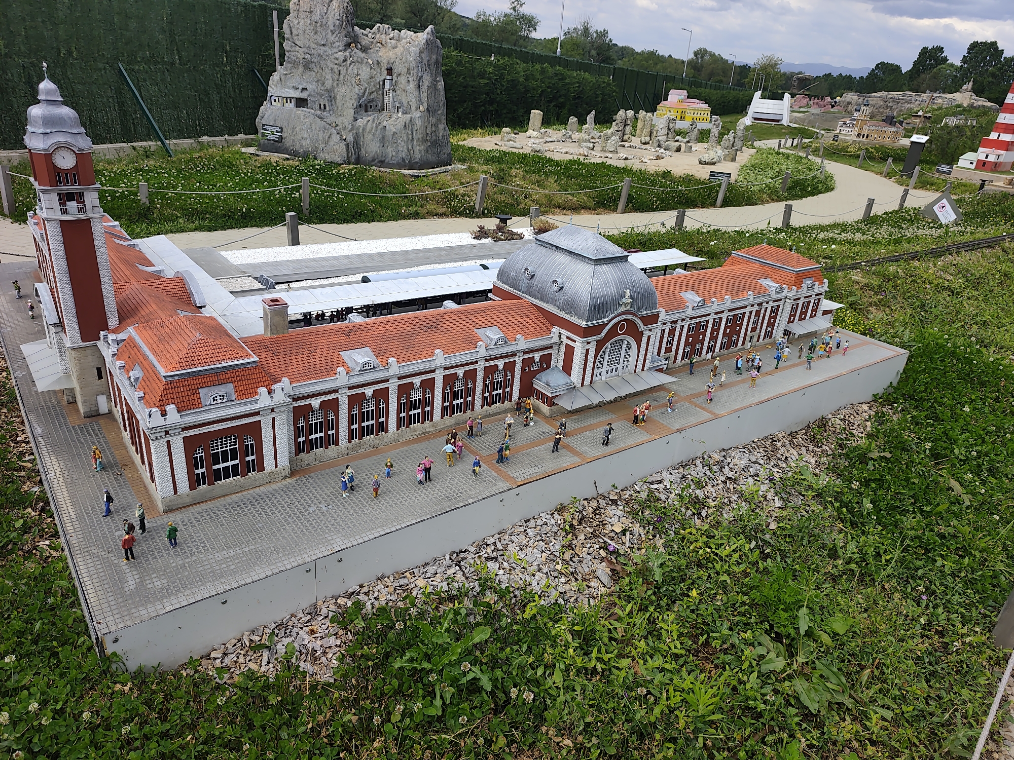 The image shows a miniature replica of a railway station located in Mini Bulgaria Park, Veliko Tarnovo, Bulgaria. The model is intricately detailed, featuring a red and white building with a clock tower and a domed roof. Tiny figures are scattered around the station, representing people walking and interacting. The station is set within a landscaped environment with grass, trees, and other miniature landmarks.