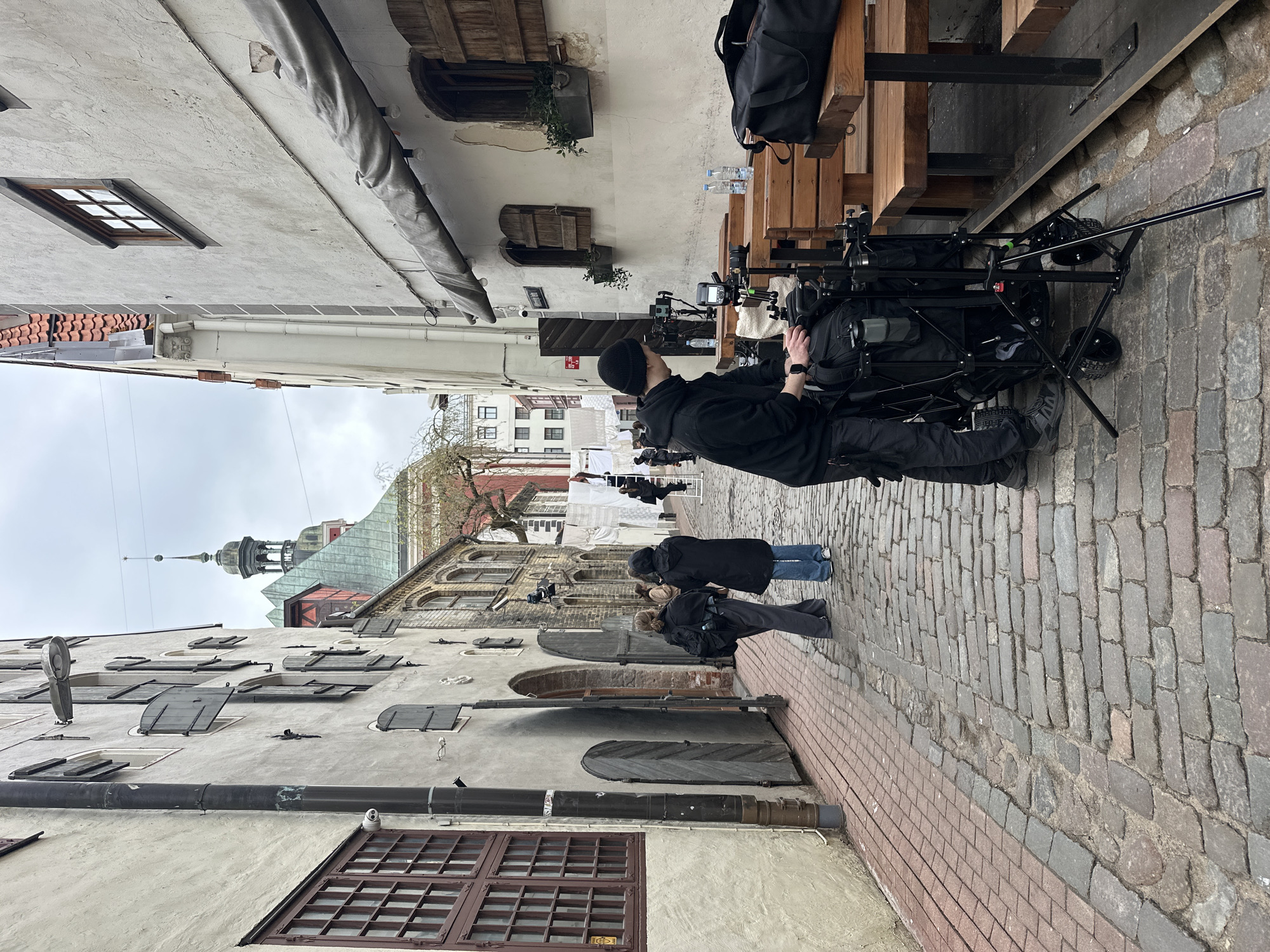 The image depicts a narrow cobblestone street in Riga, Latvia. On the left, a multi-story building with light-colored walls, dark window frames, and various architectural details lines the street. Pipes run along the building's exterior. On the right, tables and benches are positioned along the street. A person wearing black clothing and a beanie is operating a camera mounted on a wheeled platform. Further down the street, two people are crouched down, appearing to be observing or filming something. In the background, a church steeple is visible. Laundry is hung in the distance, suggesting residential activity. The sky is overcast, casting a soft light on the scene.