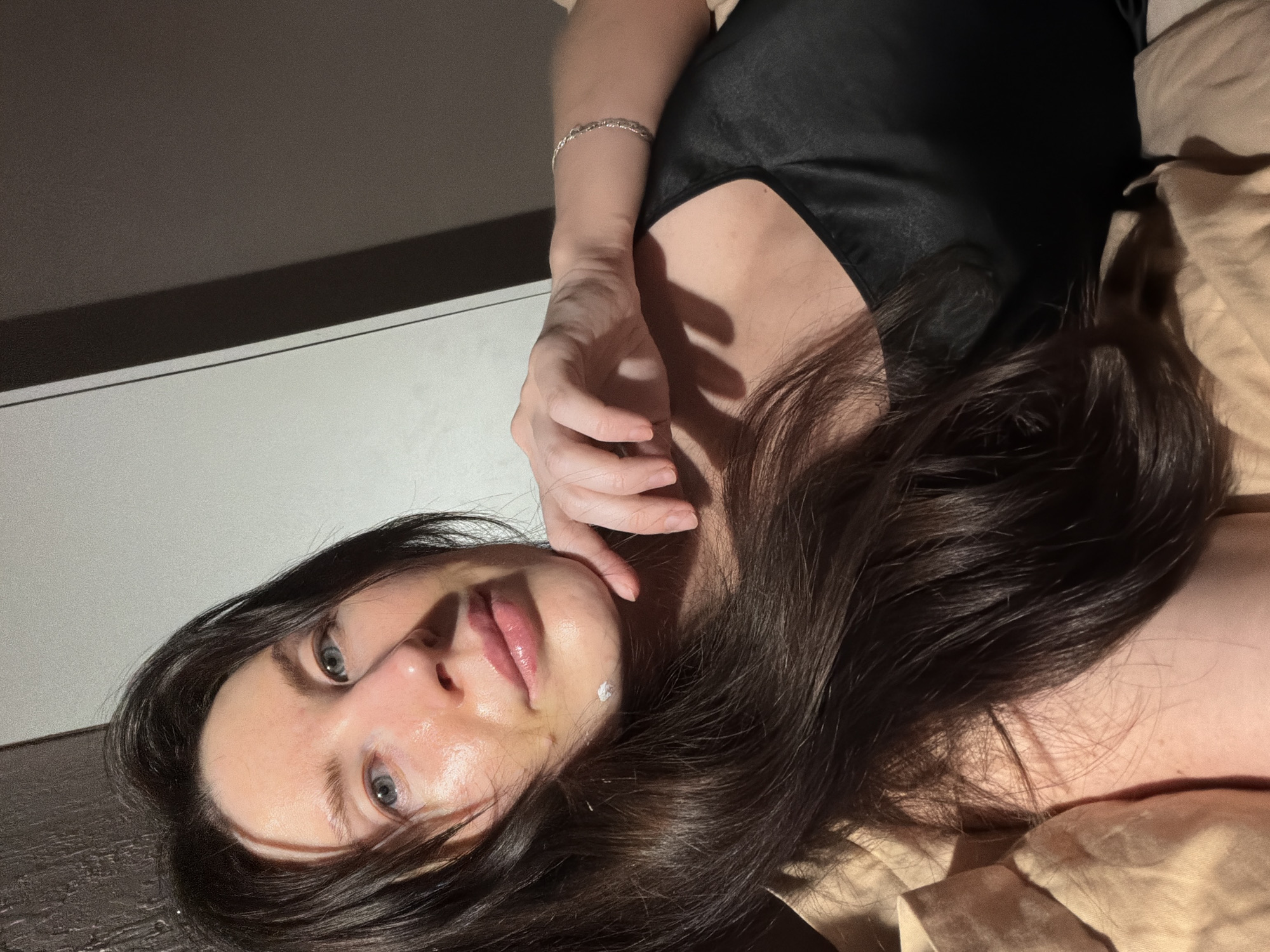 The image displays a close-up, self-portrait of an adult female in an indoor setting. The precise location within Perm, Russia, is undetermined from the visual information. The subject, possessing long, dark brown hair and blue-grey eyes, is positioned from the chest up, partially reclined against beige, rumpled bedding. She wears a black, sleeveless top, possibly satin. A silver-colored chain-link bracelet is present on her right wrist. Her left hand is raised, with fingers slightly curled, touching her chin/jaw area. A distinct white, opaque substance, consistent with a cream or topical application, is visible on the left side of her chin. The background consists of a light-colored vertical panel on the left, separated by a dark vertical line from a darker, solid-colored panel on the right. Illumination appears to be strong and directional, highlighting the right side of the subject's face and upper body, and casting shadows on the left. The subject is looking directly into the camera.