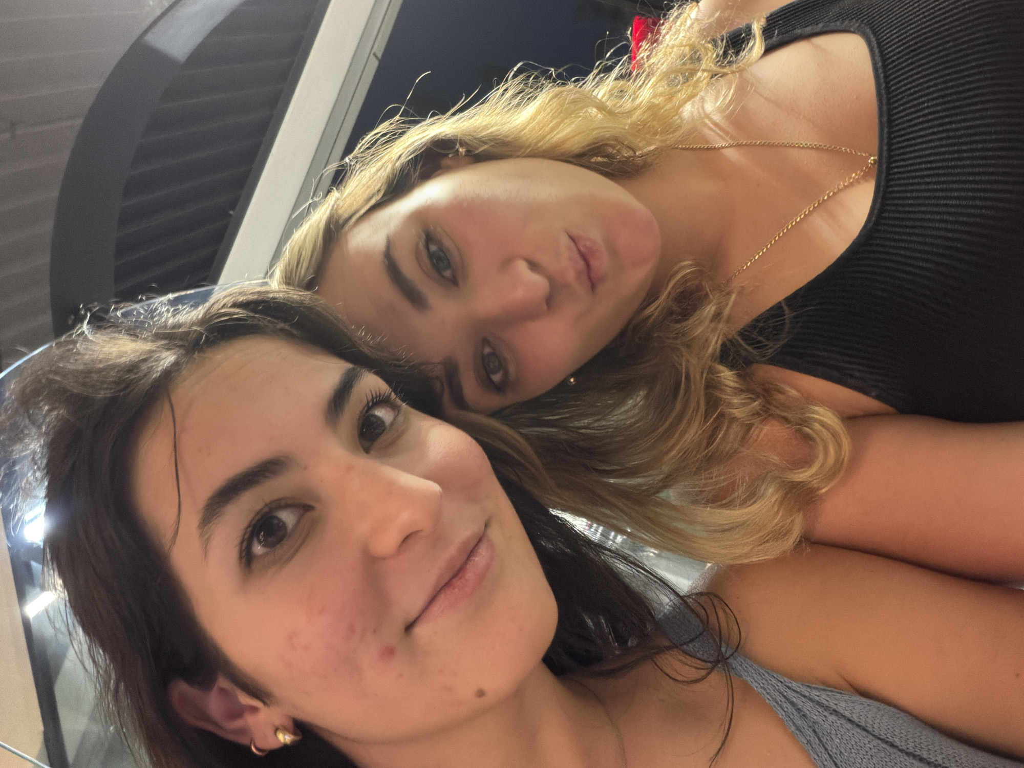 This close-up, angled selfie features two young women in an indoor setting, likely during the evening or night.
The woman in the foreground, positioned in the lower-left, has dark brown hair and eyes. She is wearing a light grey or blue knitted top. A small, gold-toned hoop earring, with a decorative spike, is visible on her left ear. Her expression is a subtle, closed-mouth smile, and she has a few minor blemishes and freckles visible on her cheek.
Behind and slightly above her, the second woman has wavy blonde hair and lighter eyes. She is wearing a black ribbed top and a delicate gold chain necklace. Her expression is a playful pout, with her lips slightly puckered towards the camera.
The background is dark and out of focus, suggesting an interior space with artificial lighting. Indistinct shapes, possibly parts of a wall or architectural features with horizontal lines, are visible behind the women. No specific location cues are discernible within the image itself. The overall mood is casual and friendly. No text is visible in the image.