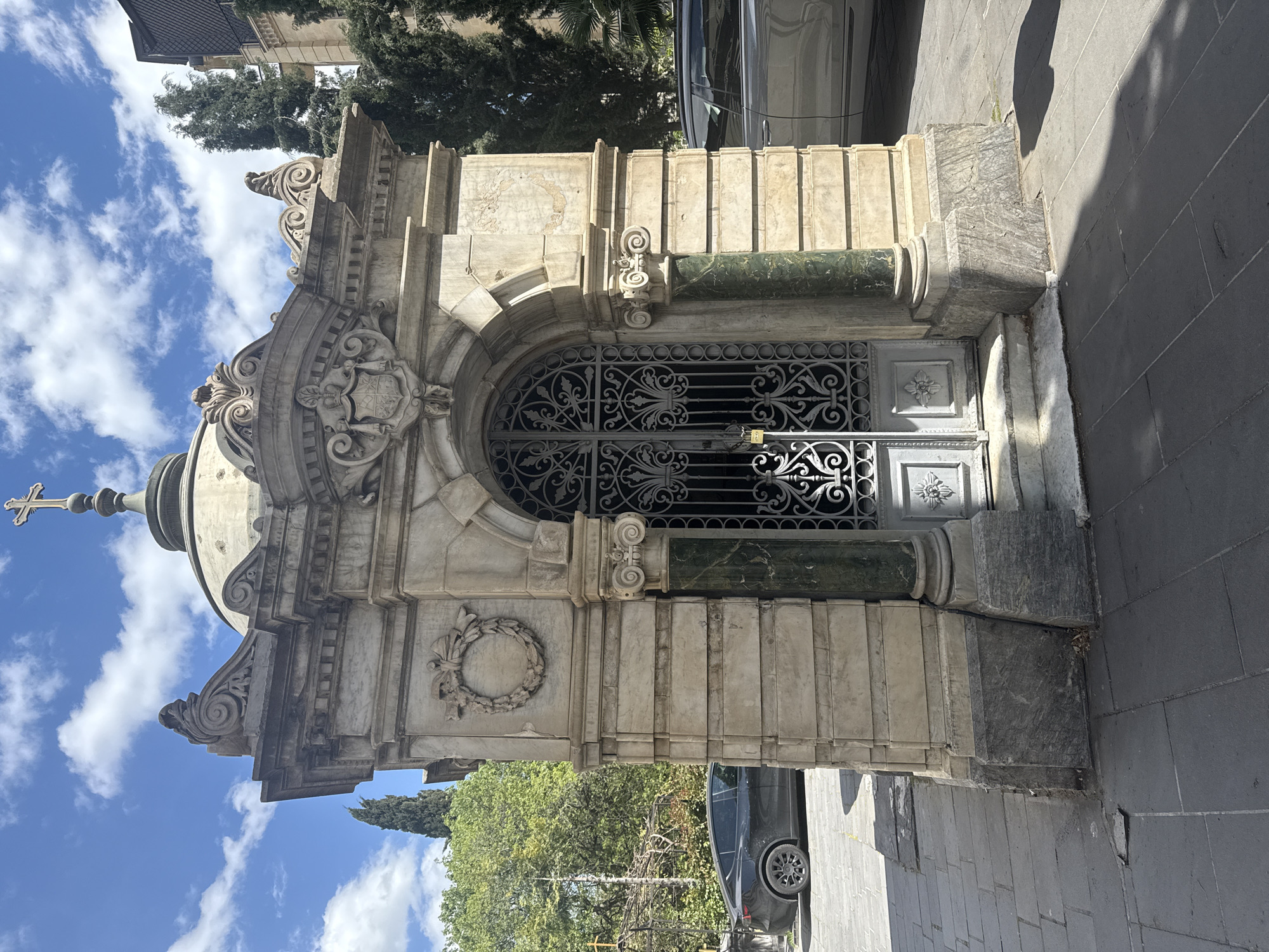 A vertical, medium shot depicts an ornate, stone structure located in Tbilisi, Georgia, likely a mausoleum or entrance to a cemetery. The architectural style appears historical, with elements like a wrought-iron gate, marble or limestone facade, decorative carvings including a possible coat of arms, and a small dome topped with a cross. The structure is built on a sidewalk with grey paving stones, and a portion of a car is visible on the right side of the frame. Trees are visible in the background and blue sky with white clouds at the top of the frame. The style of the structure seems European in influence. The lighting is bright and appears to be taken during the daytime.