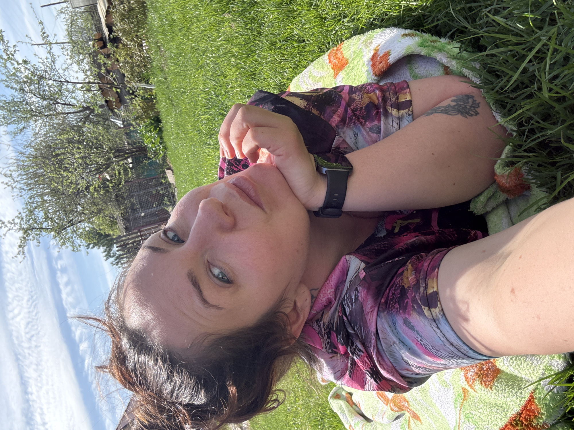 A woman lies on the grass outdoors in Chaikovsky, Russia. She is wearing a floral-patterned top and a black smartwatch on her left wrist. Part of a patterned towel lies beneath her. There is a tattoo on her left arm. The environment includes green grass and trees. The sky is partially visible in the background.