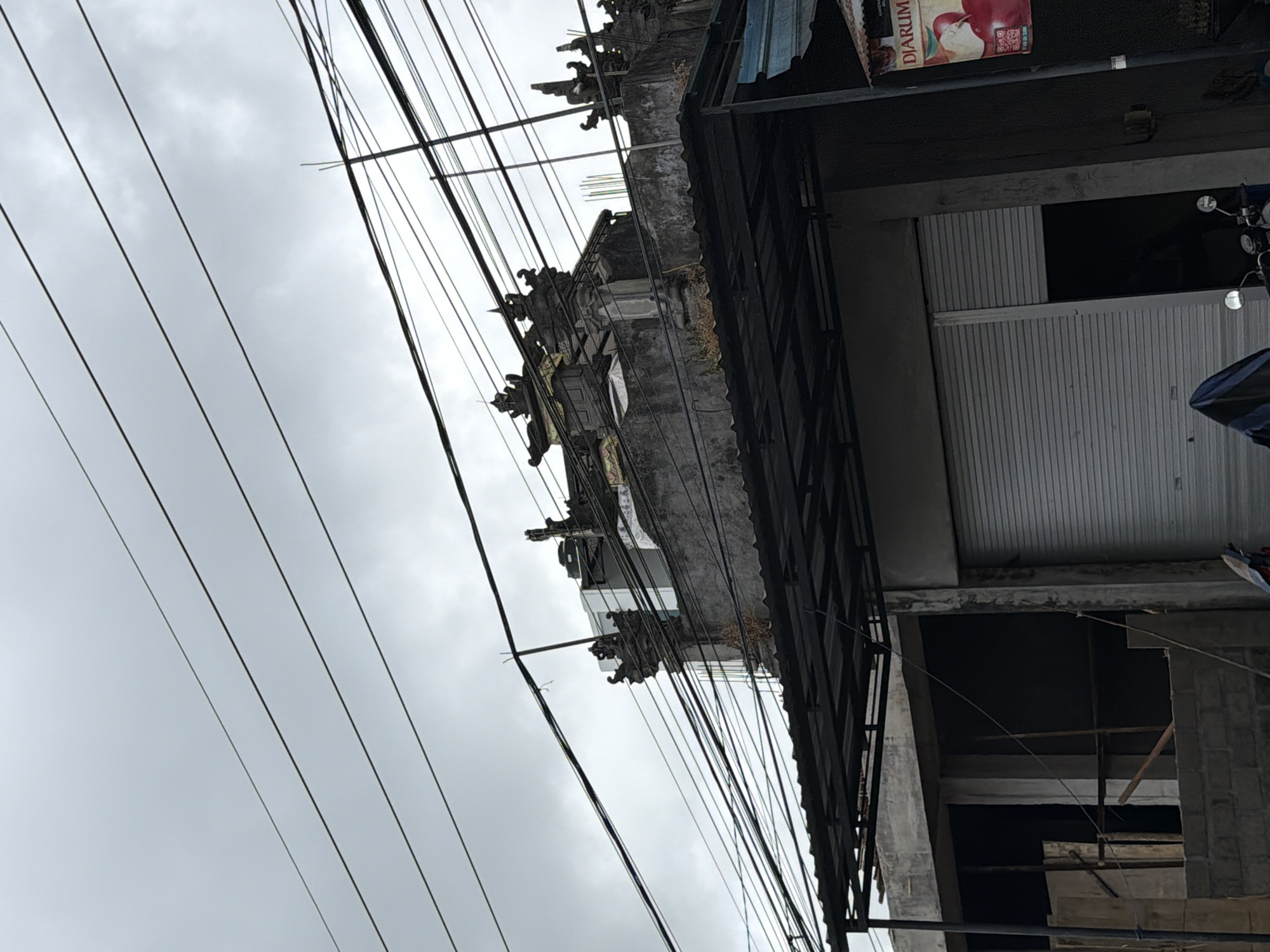 The image depicts the rooftop and upper facade of a building in Kintamani, Indonesia, against a cloudy sky. The building features traditional Balinese architectural elements on its roof, with ornate stone carvings and tiered structures. A black metal awning extends from the building's upper floor, covering a portion of the facade below. The lower part of the building shows construction in progress, with exposed brickwork and unfinished walls. A grey roller door is visible, suggesting a commercial or storage space. Numerous electrical wires are strung across the scene, obscuring the sky in many areas. A red and white sign with text that appears to be in Indonesian, possibly advertising a product, is attached to the right side of the building. The scene is devoid of any people or overt activity, conveying a sense of quietude.